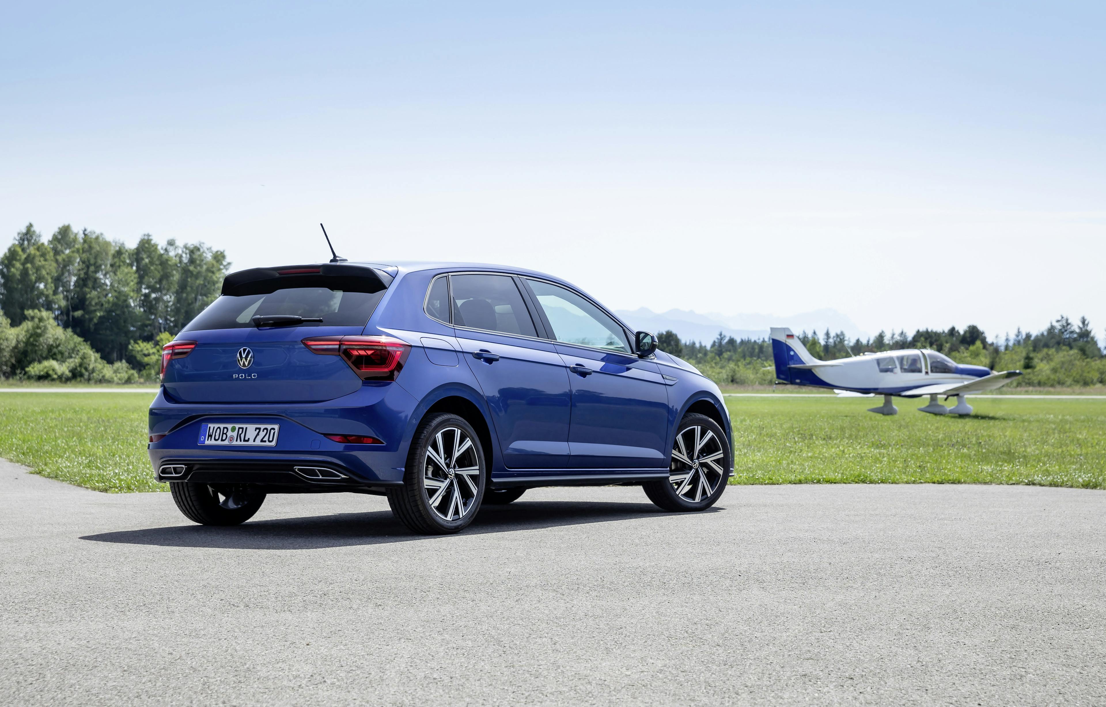 Ein blauer VW Polo steht auf einem Parkplatz neben einer Wiese mit einem Kleinflugzeug im Hintergrund.