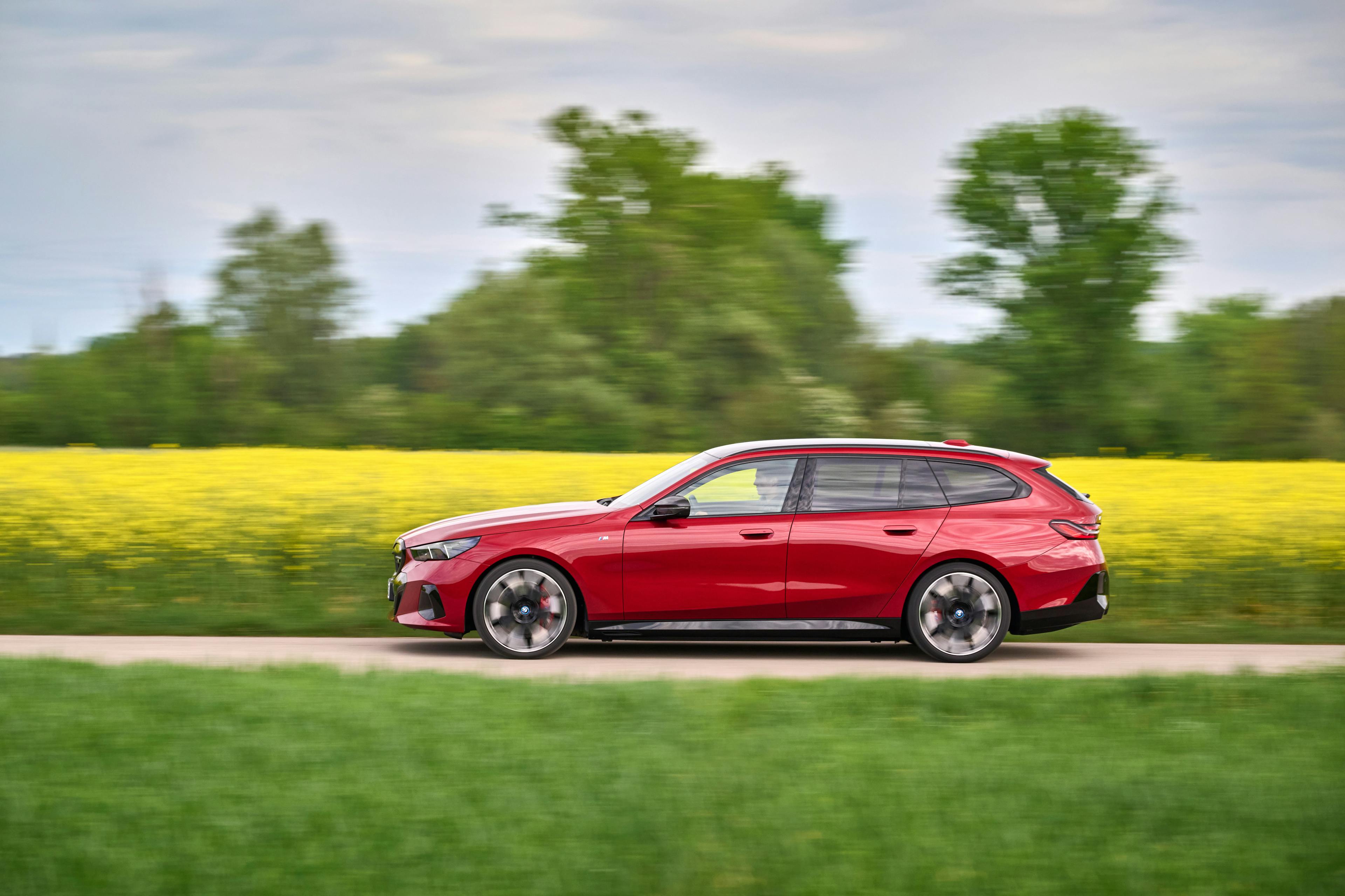  Dynamische Seitenansicht eines roten BMW i5 M60 Touring auf einer Straße mit Maisfeld im Hintergrund.