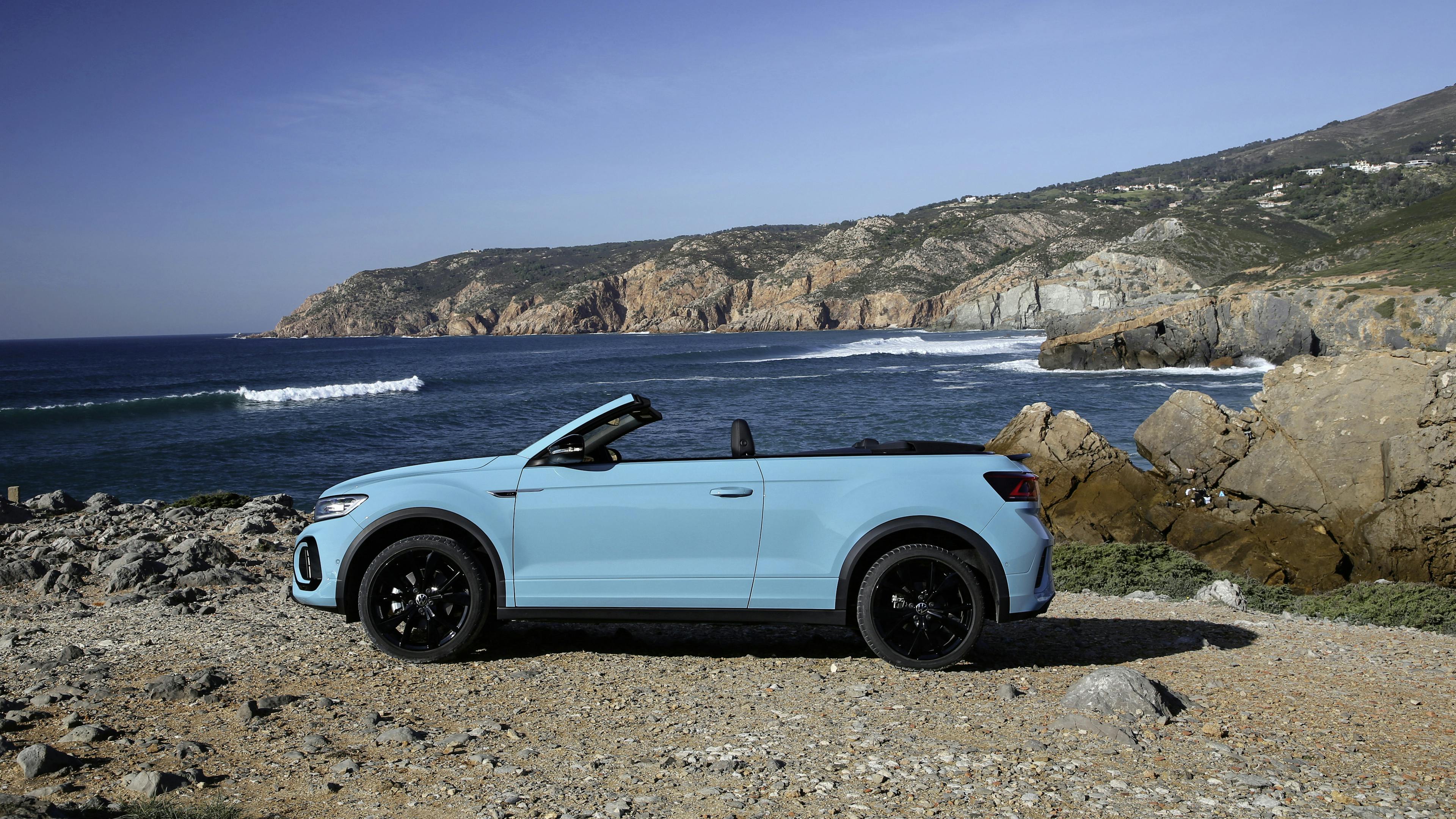 A blue T-Roc convertible stands by the sea with the top open.
