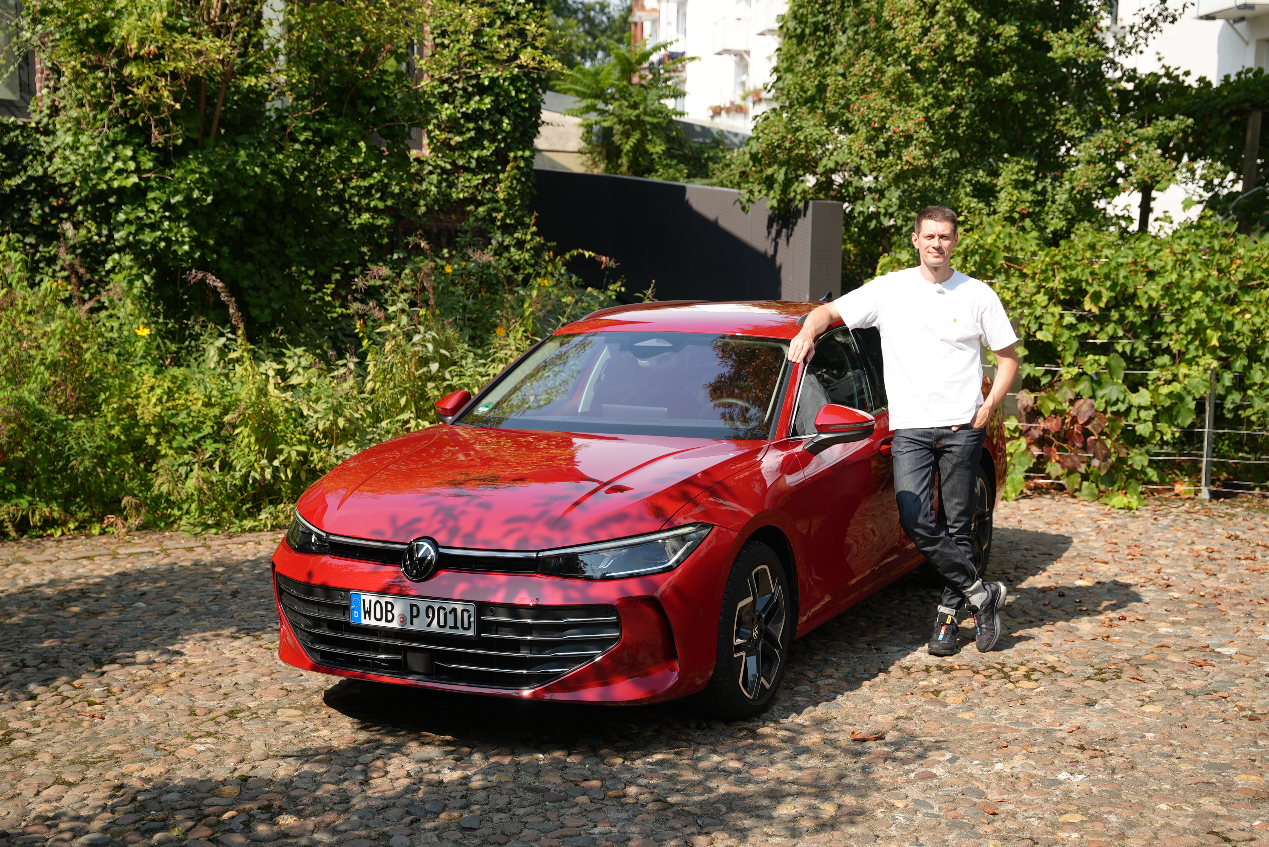 Ein roter VW Passat B9 auf Kopfsteinpflasterplatz mit Klinkergebäude im Hintergrund und dem Redakteur am Auto lehnend.