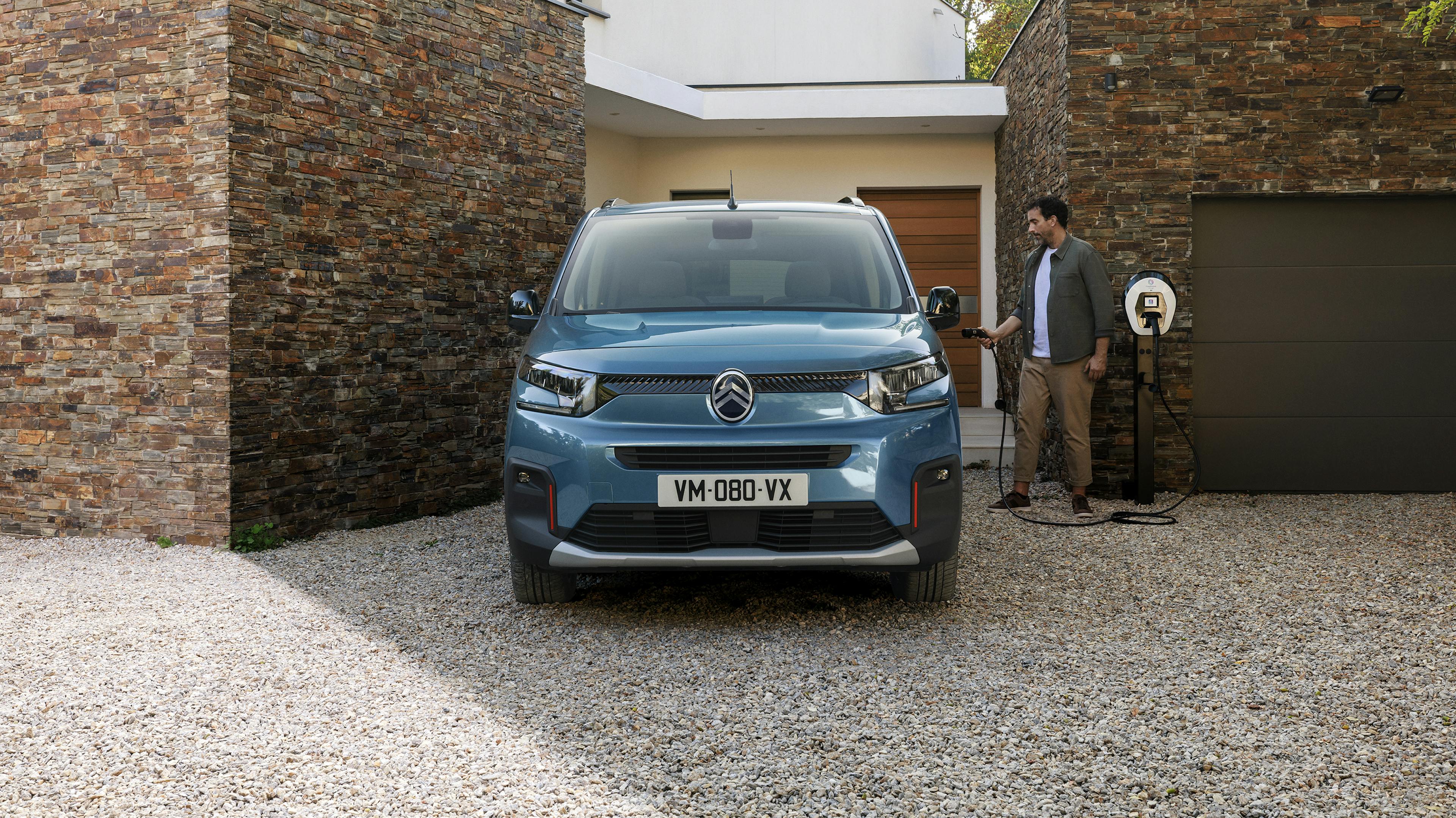 A blue Citroën van stands in front of a modern house while a man holds a charging cable in his hand.