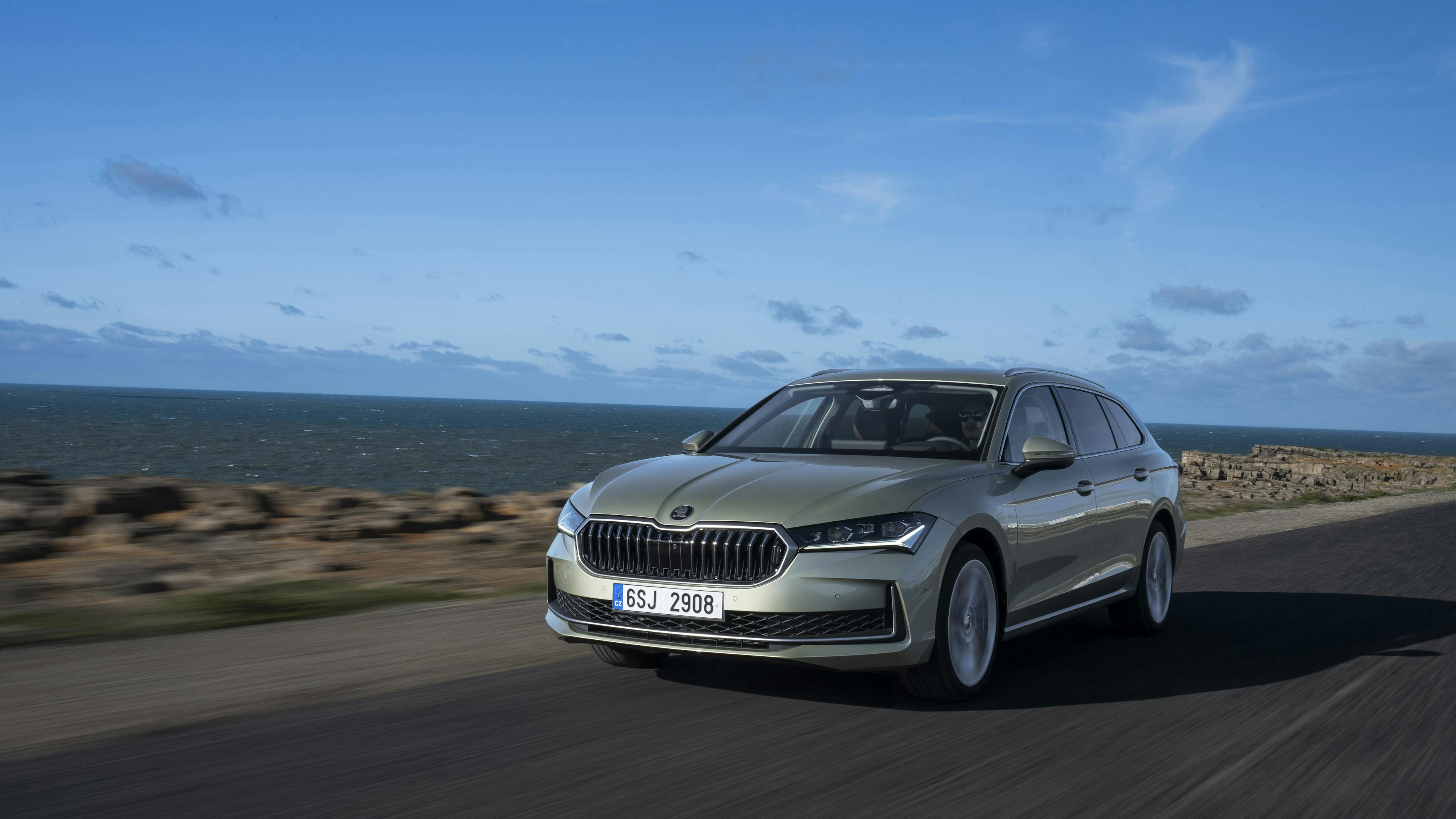 The picture shows a silver Å koda Superb Combi driving on a coastal road.