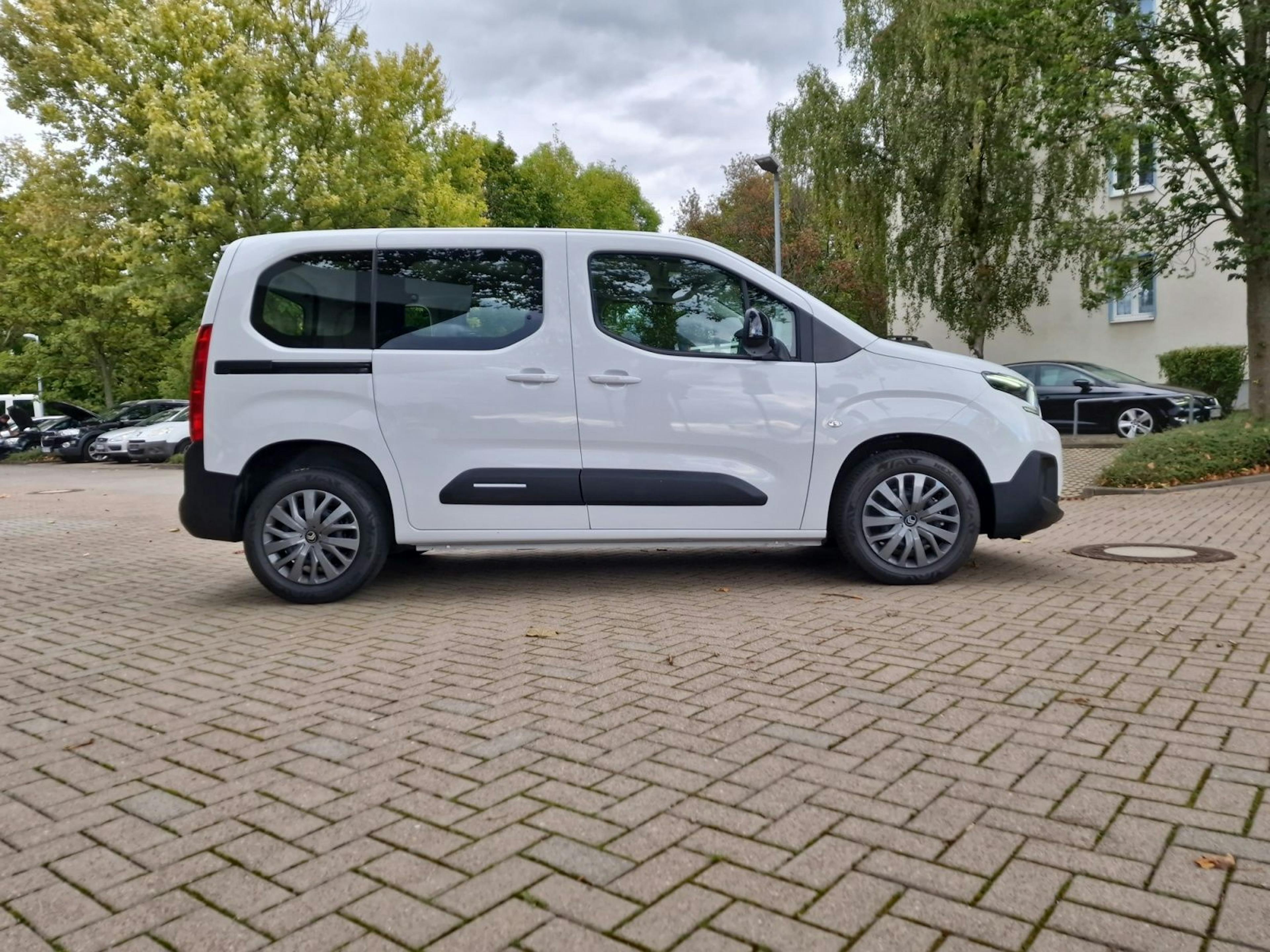 Weißer Citroën Berlingo, seitlich von rechts fotografiert, geparkt auf einem gepflasterten Parkplatz mit Bäumen und Wohnhäusern im Hintergrund.