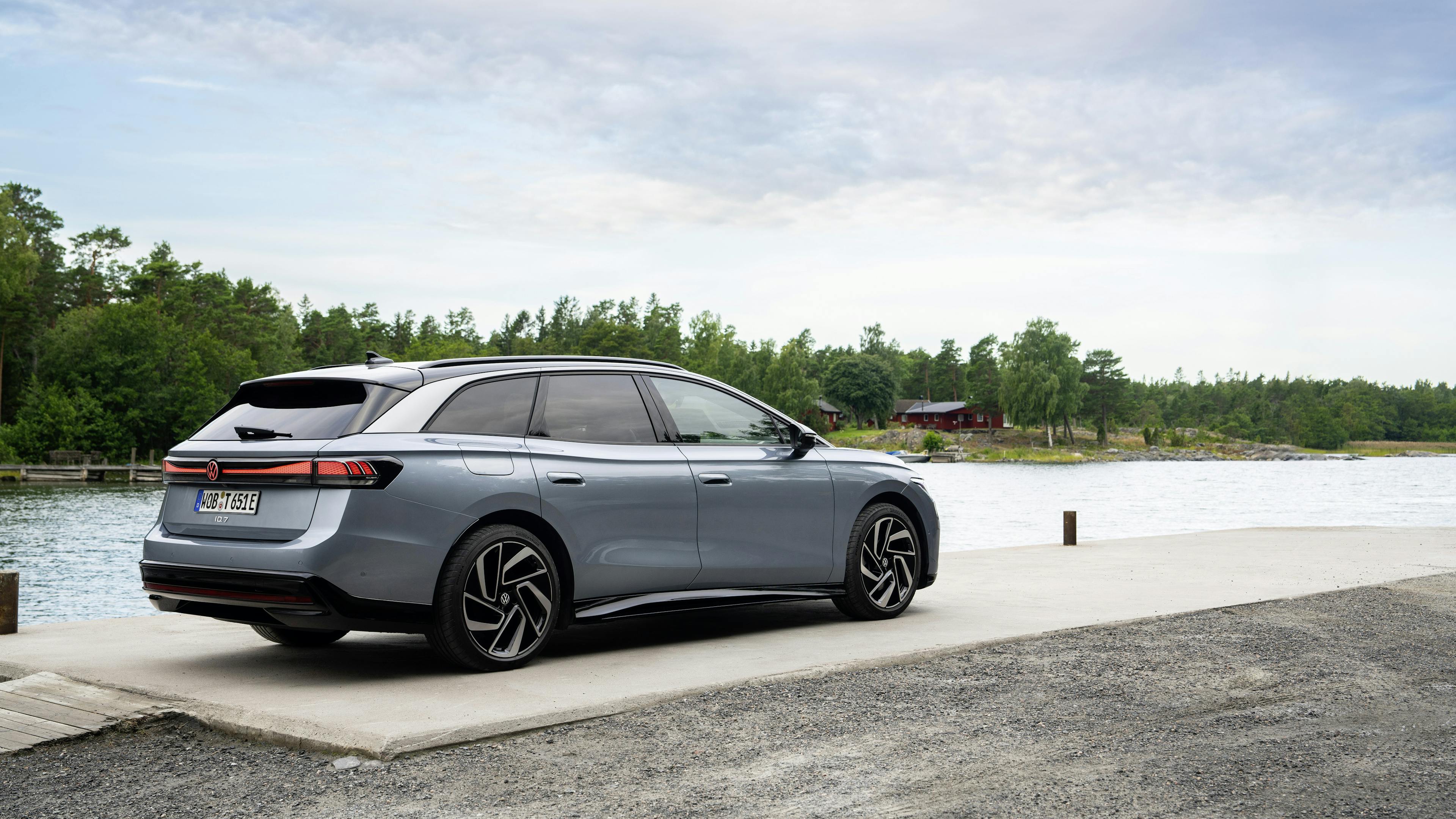 Silver VW ID.7 Tourer, photographed from diagonally behind, parked on a lakeshore with a forest and a wooden house in the background.