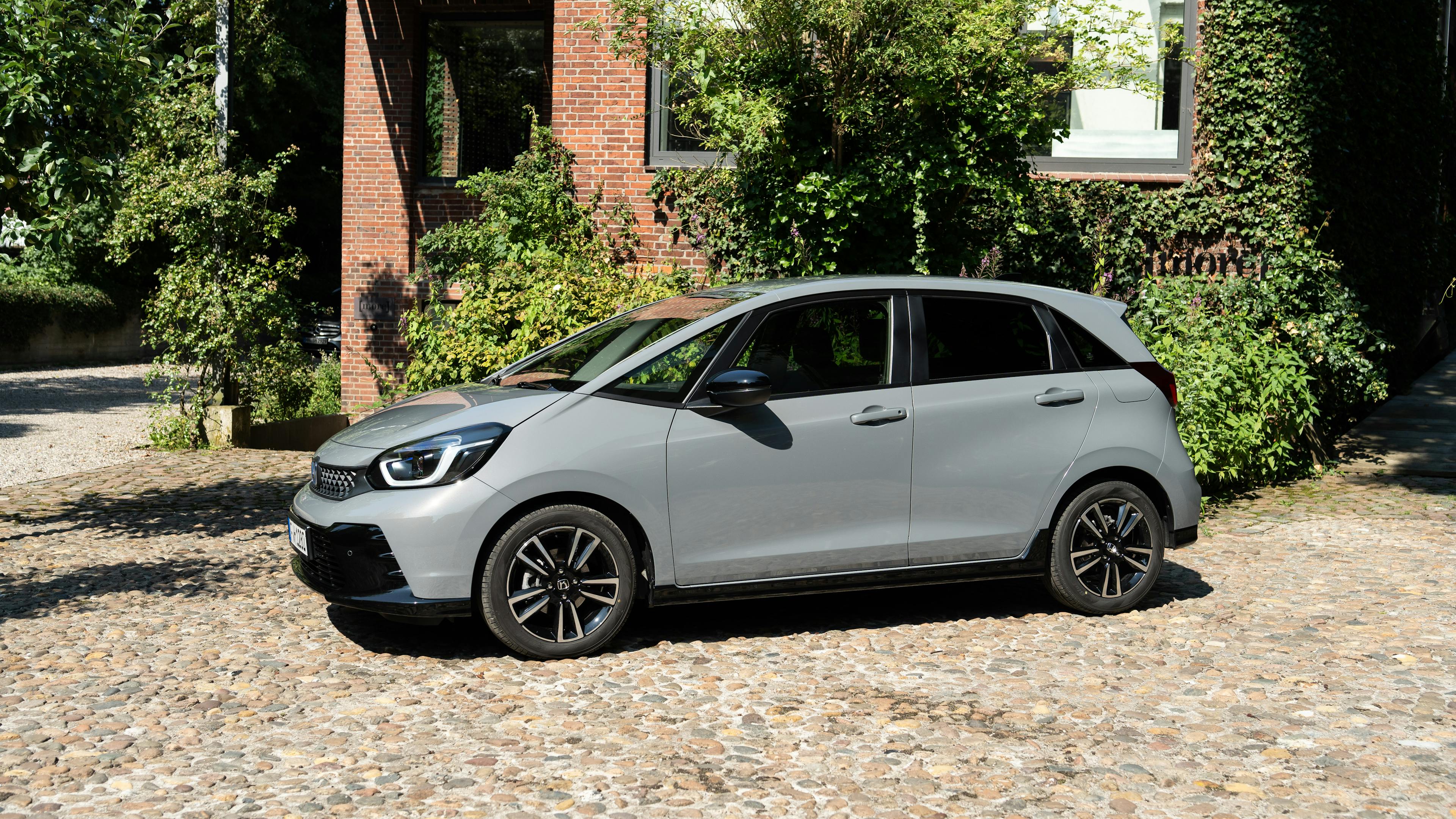 A gray Honda Jazz is parked in a cobbled square in front of a green brick building.