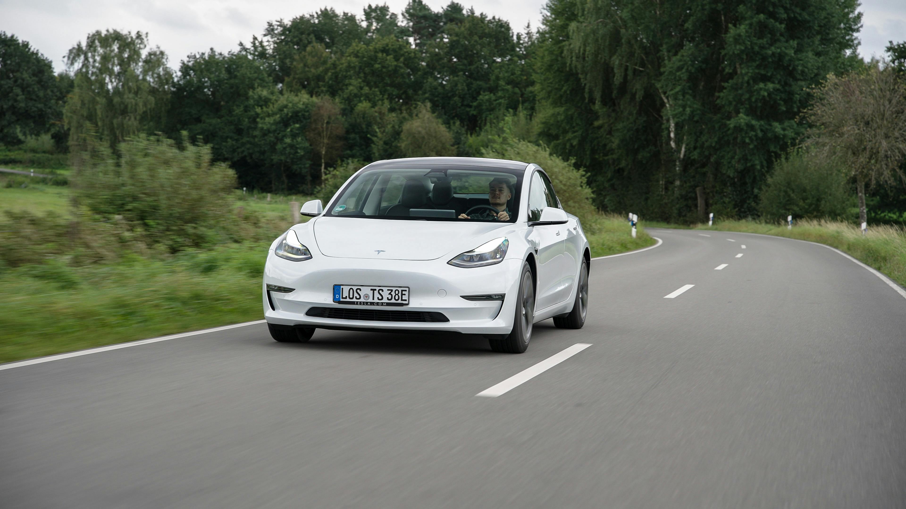 Ein weißes Tesla Model 3 während der Fahrt, von vorne auf einer Landstraße fotografiert.