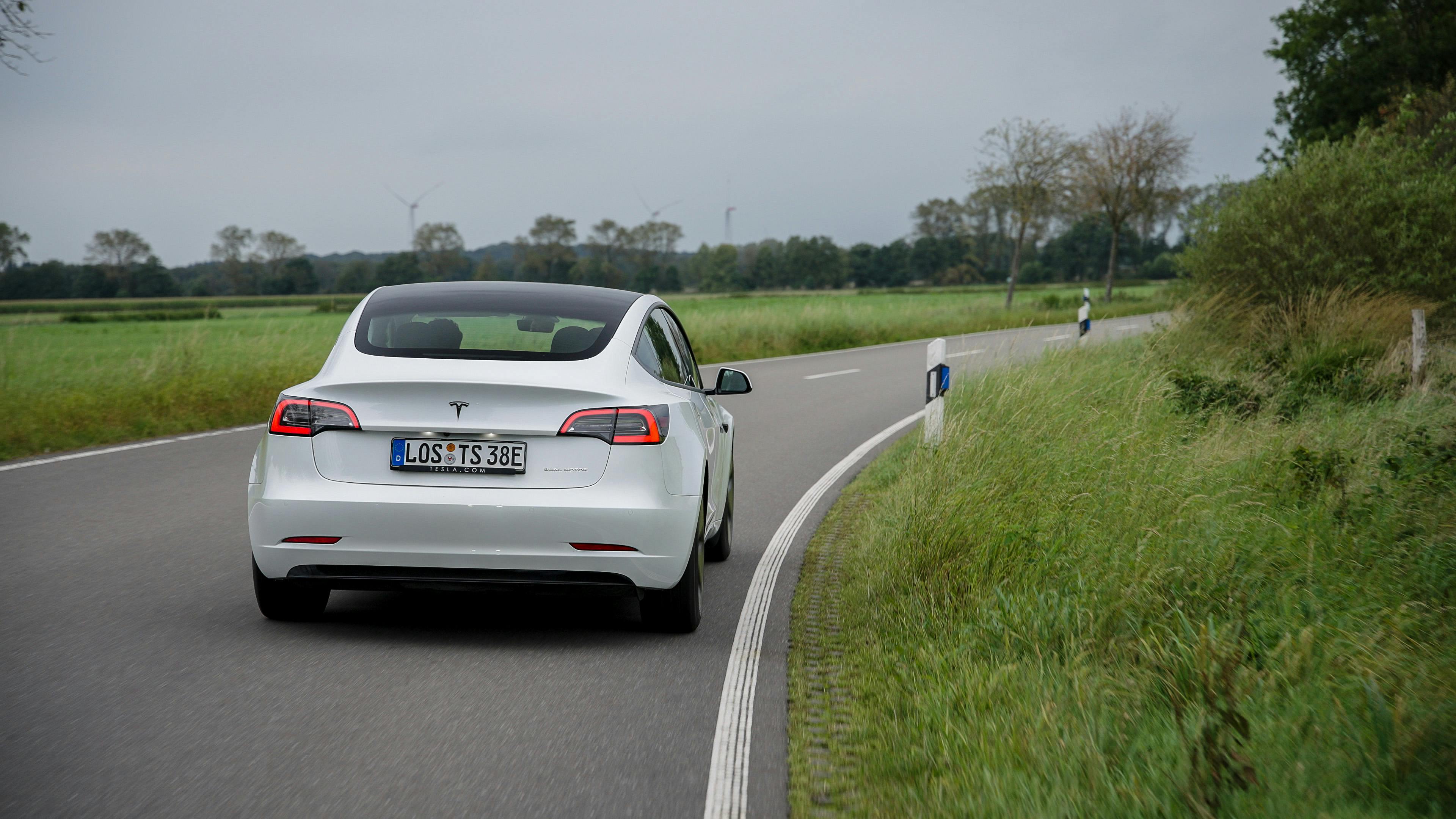 Ein weißes Tesla Model 3 während der Fahrt, von hinten auf einer Landstraße fotografiert.