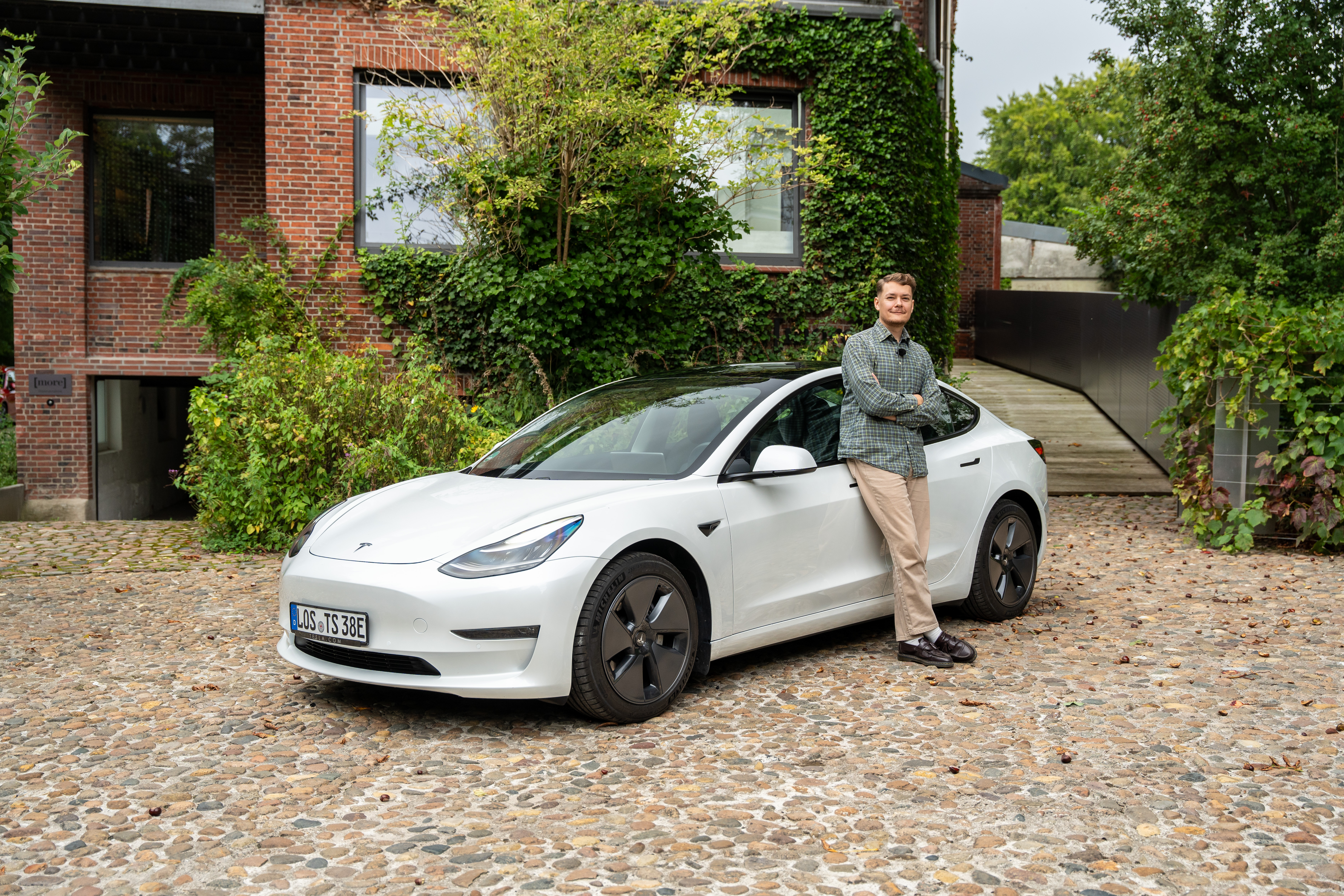 Der Redakteur lehnt an einem weißen Tesla Model 3, das schräg vor einem Backsteingebäude parkt.