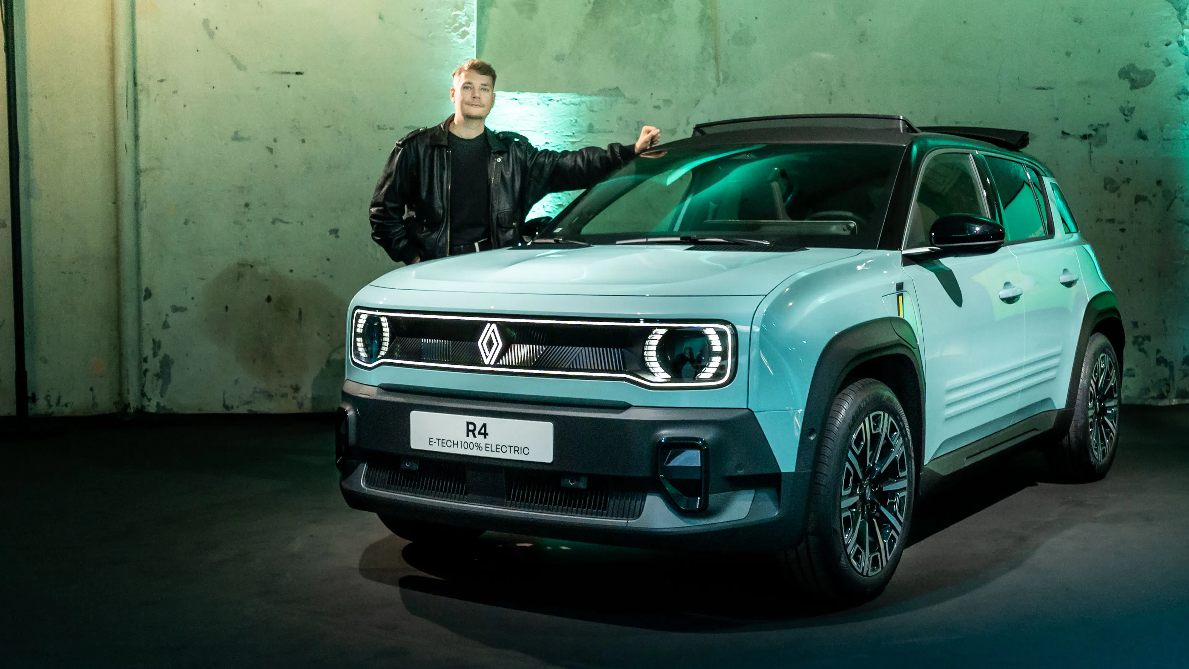 Ein Mann lehnt an einem hellblauen Renault R4 E-Tech Electric, der in einer industriellen Umgebung präsentiert wird.