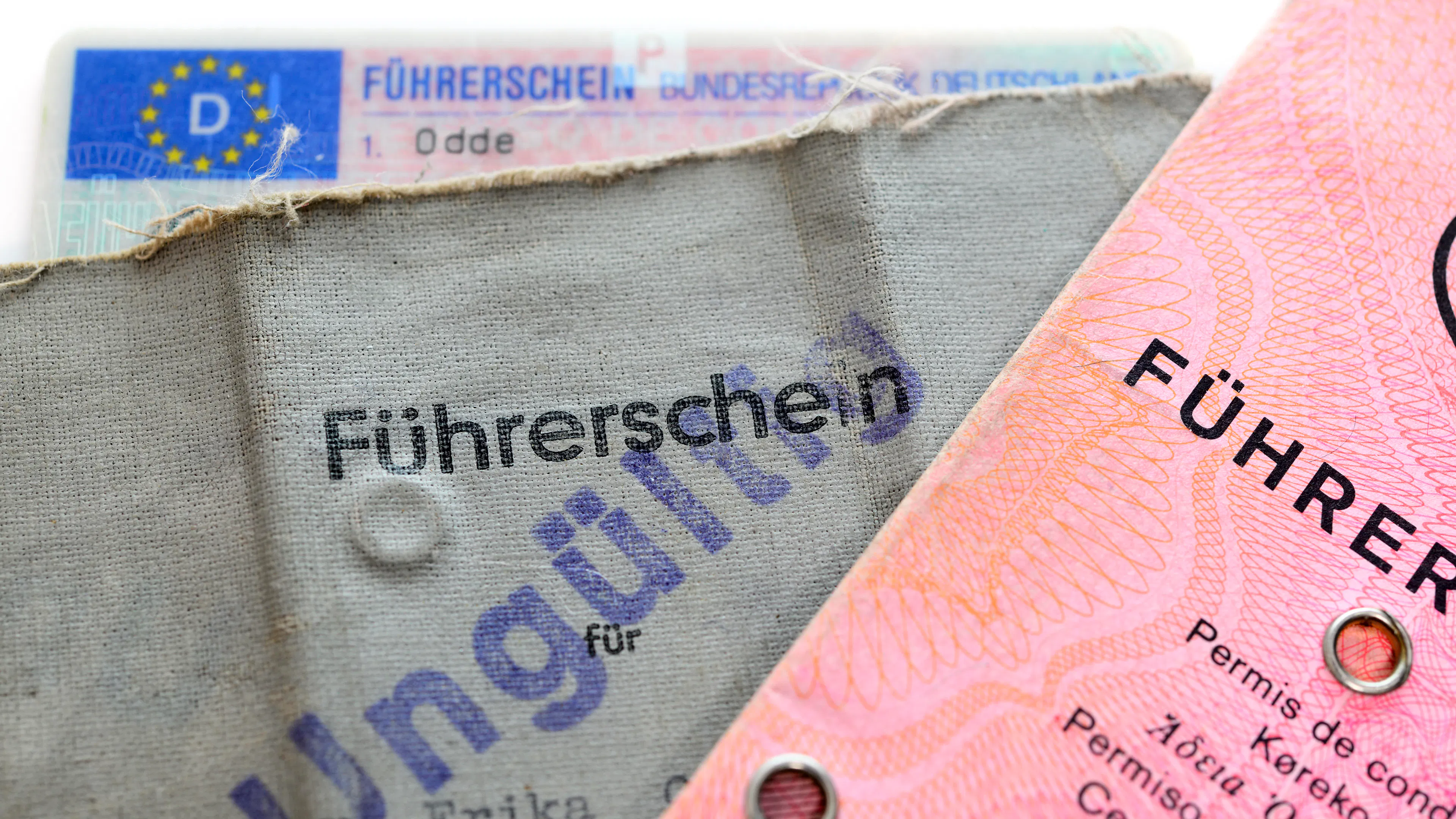 Abgebildet sind mehrere Führerscheine, darunter ein ungültig gestempelter grauer Papierführerschein und ein rosafarbener Führerschein im klassischen Design.