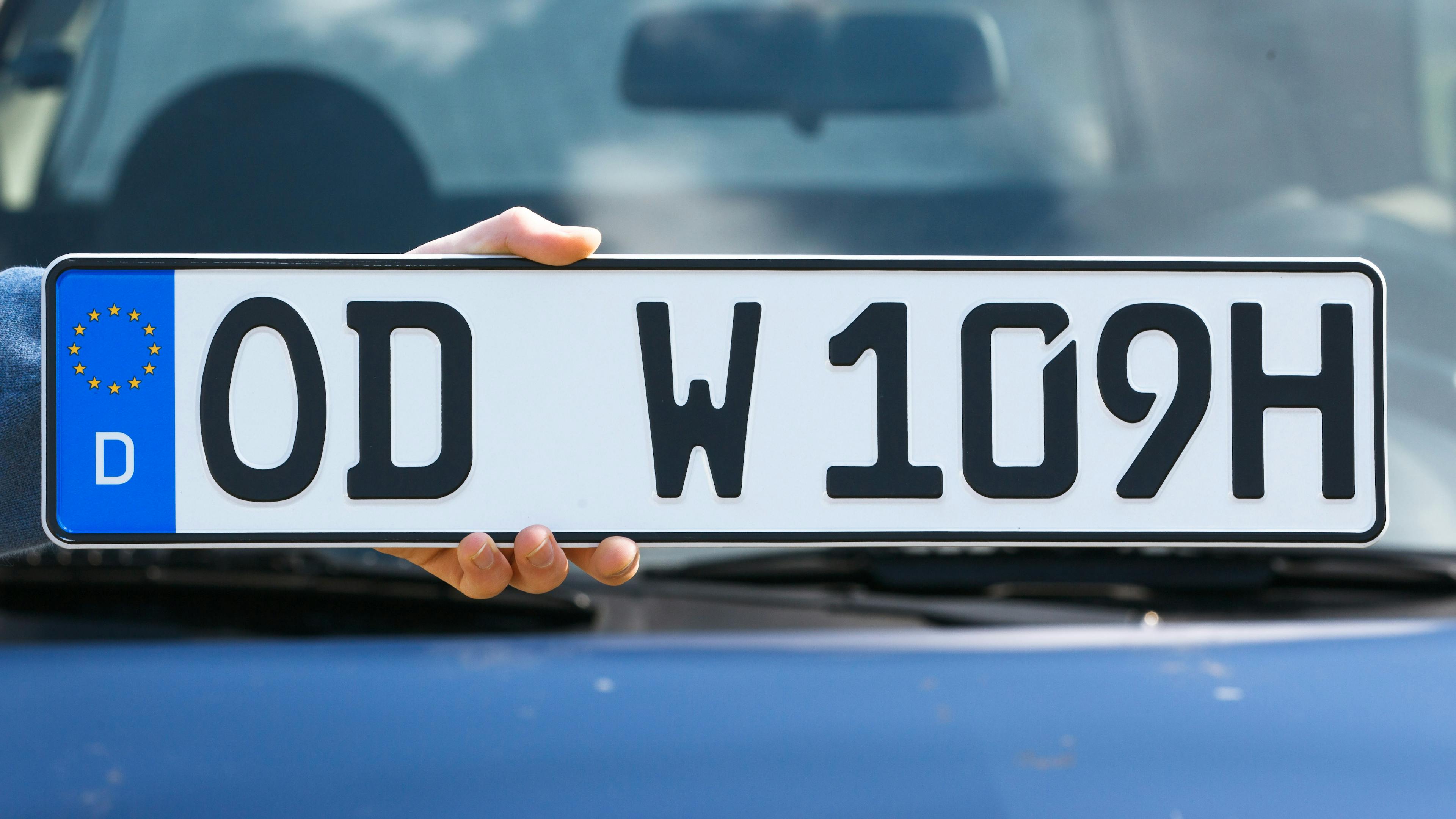 In front of a car, a person holds up a German H license plate with the inscription "OD W 109H". It marks the classic car status for vehicles aged 30 and over.