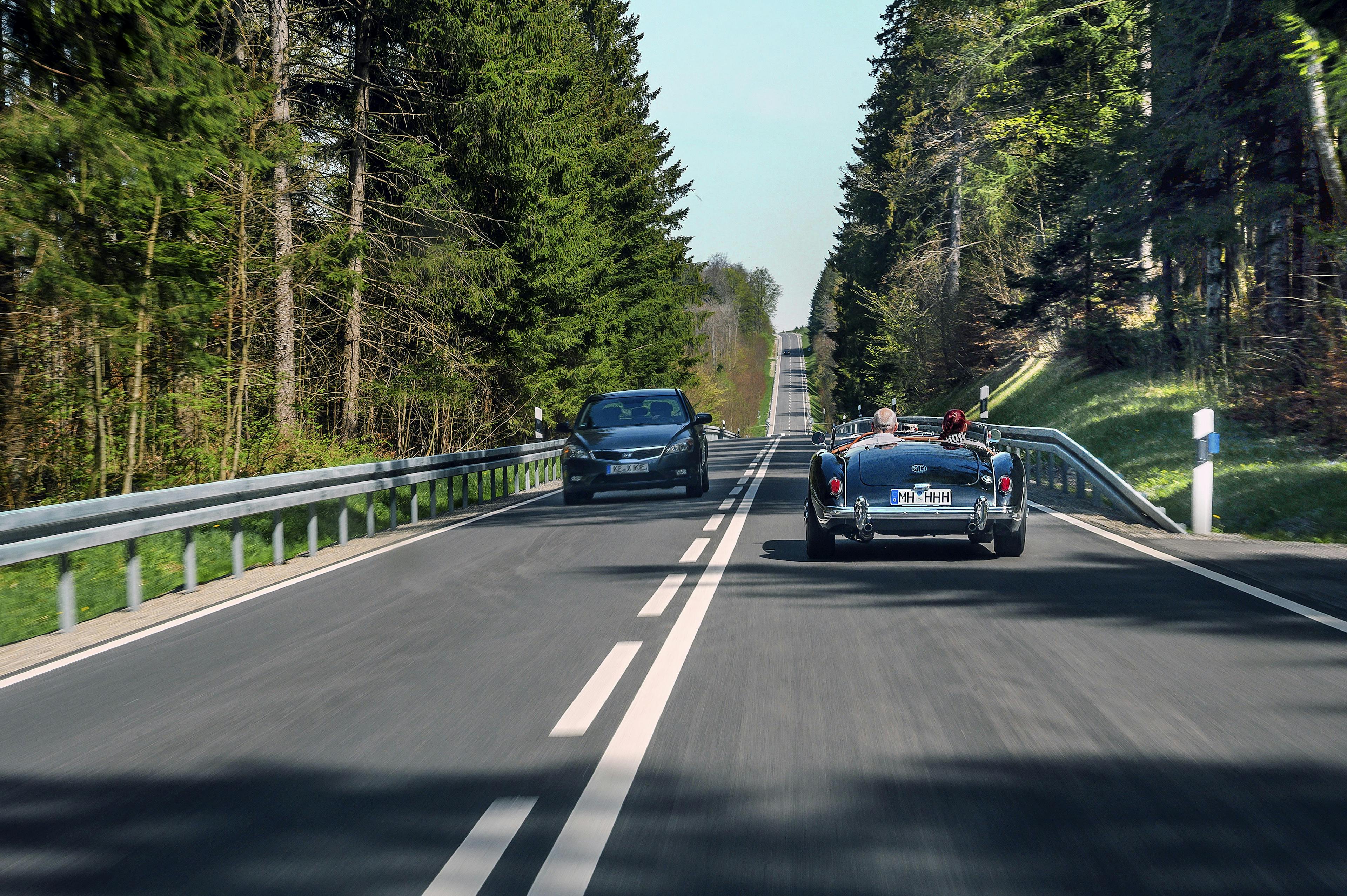 Ein blaues Oldtimer-Cabrio mit H-Kennzeichen fährt auf einer Landstraße durch eine waldreiche Umgebung. Auf der anderen Fahrbahnseite kommt dem Oldtimer ein modernes Fahrzeug entgegen.