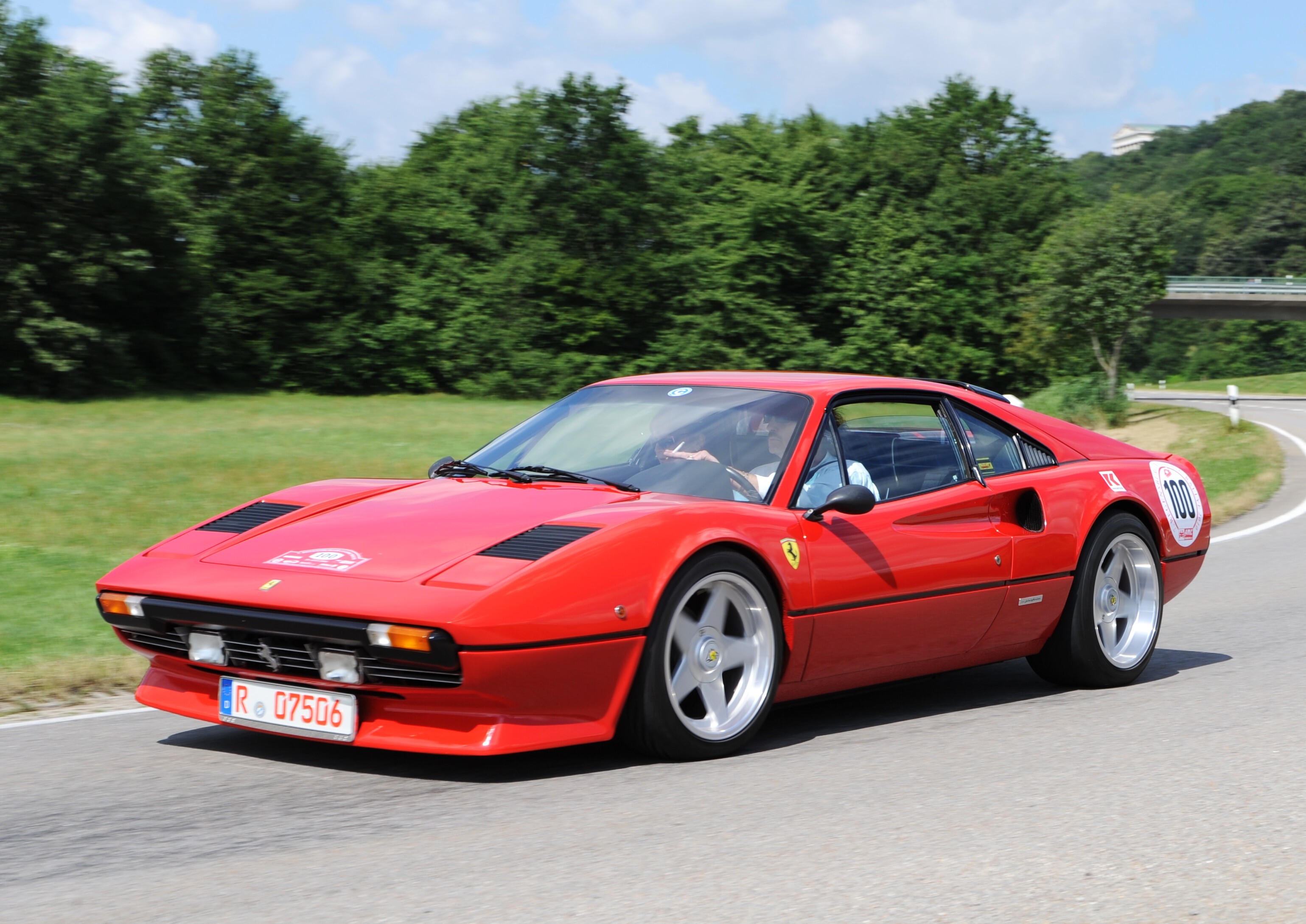 A red Ferrari classic car with a red 07 license plate drives on a country road, surrounded by green nature.