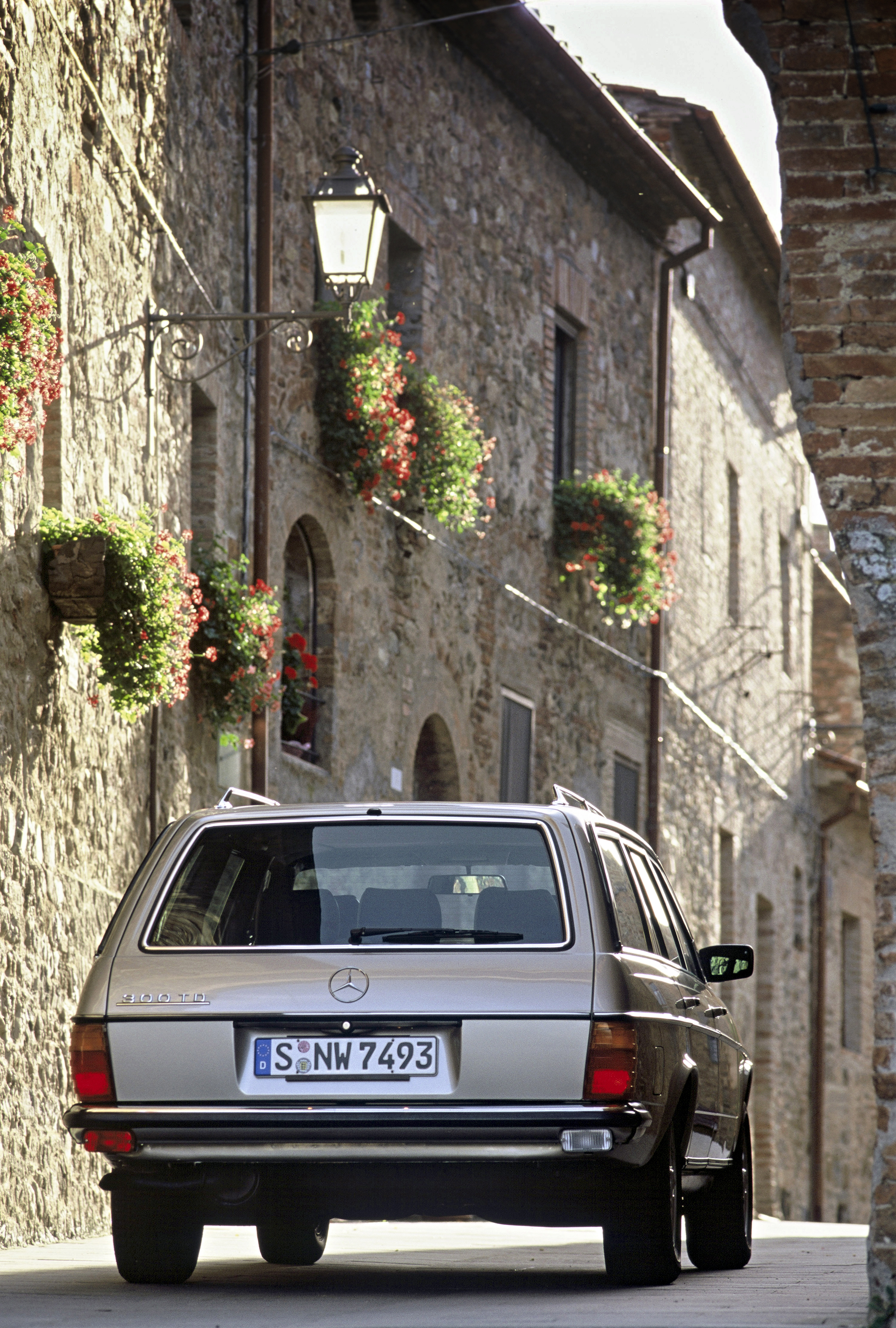 Heckansicht eines beigefarbenen Mercedes-Benz W123, der durch eine enge Gasse fährt.