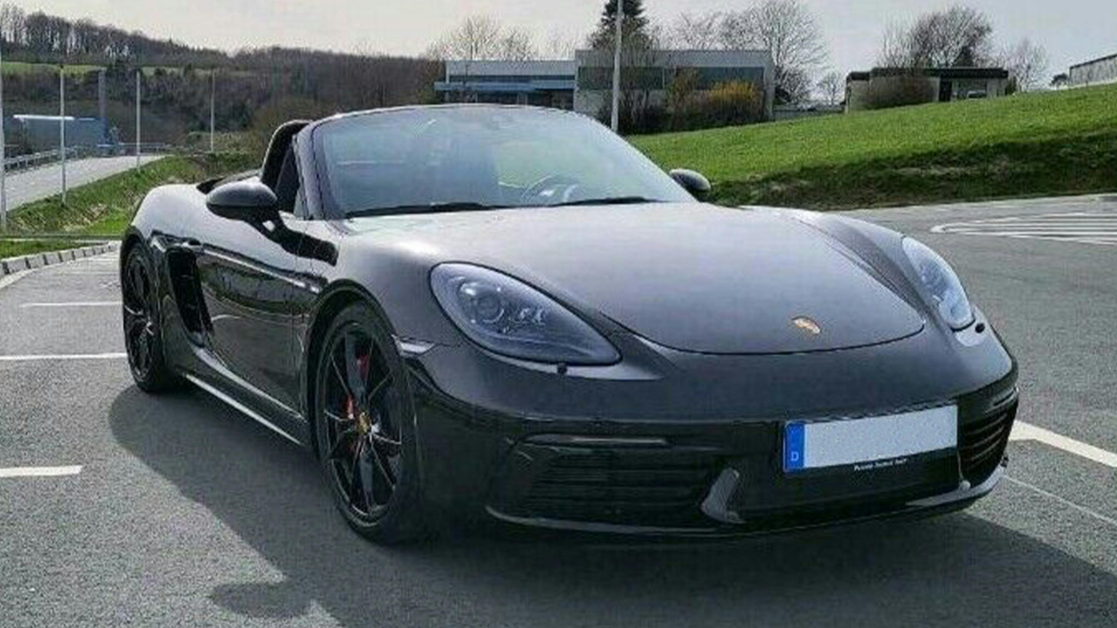 Schwarzer Porsche 718 Boxster Cabrio auf einem Parkplatz, Frontansicht. Hintergrund mit grüner Wiese, Straße und Gebäuden.