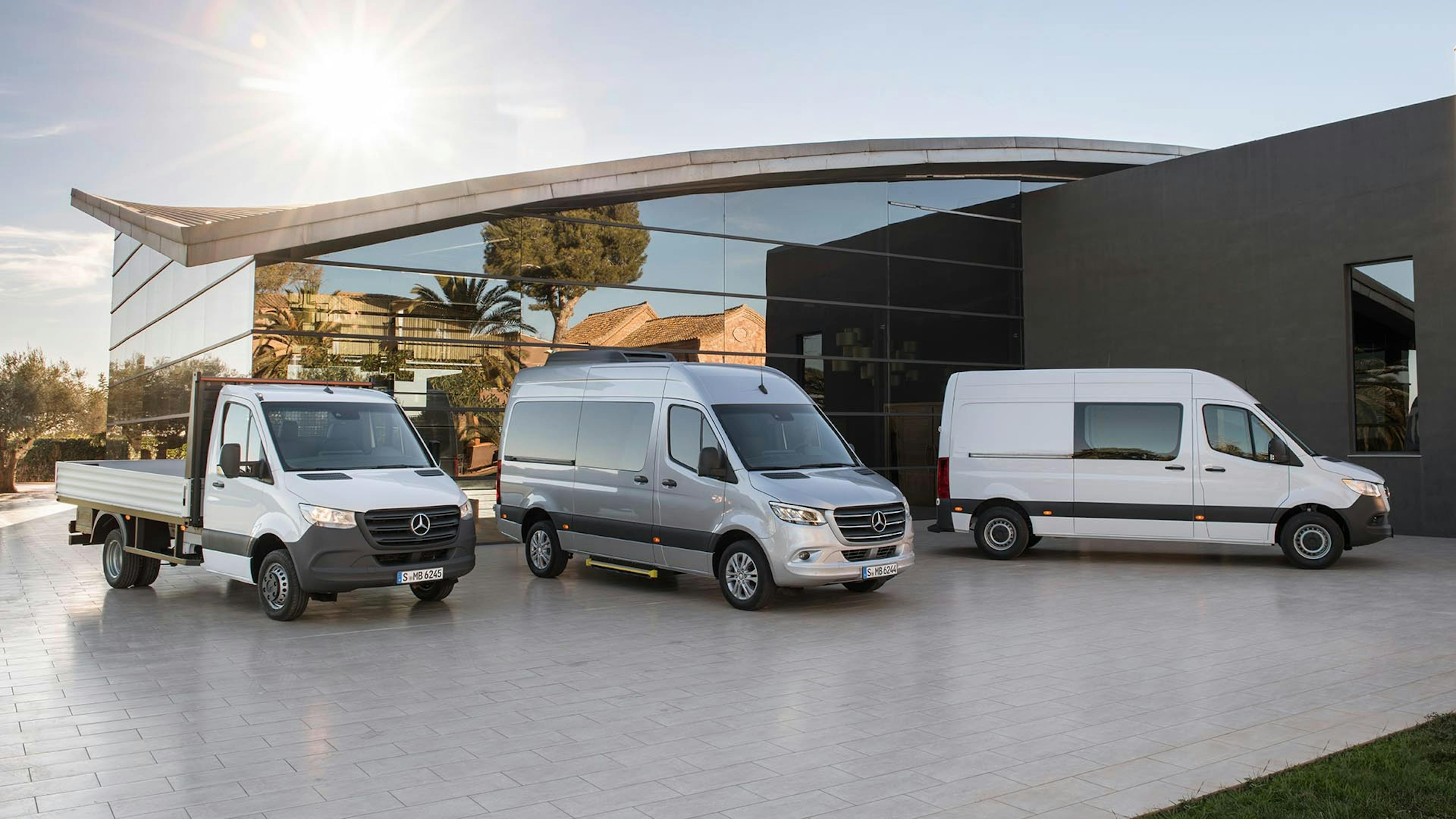 Drei Mercedes-Benz Sprinter-Modelle in unterschiedlichen Ausführungen stehen vor einem modernen Bürogebäude in der Sonne.
