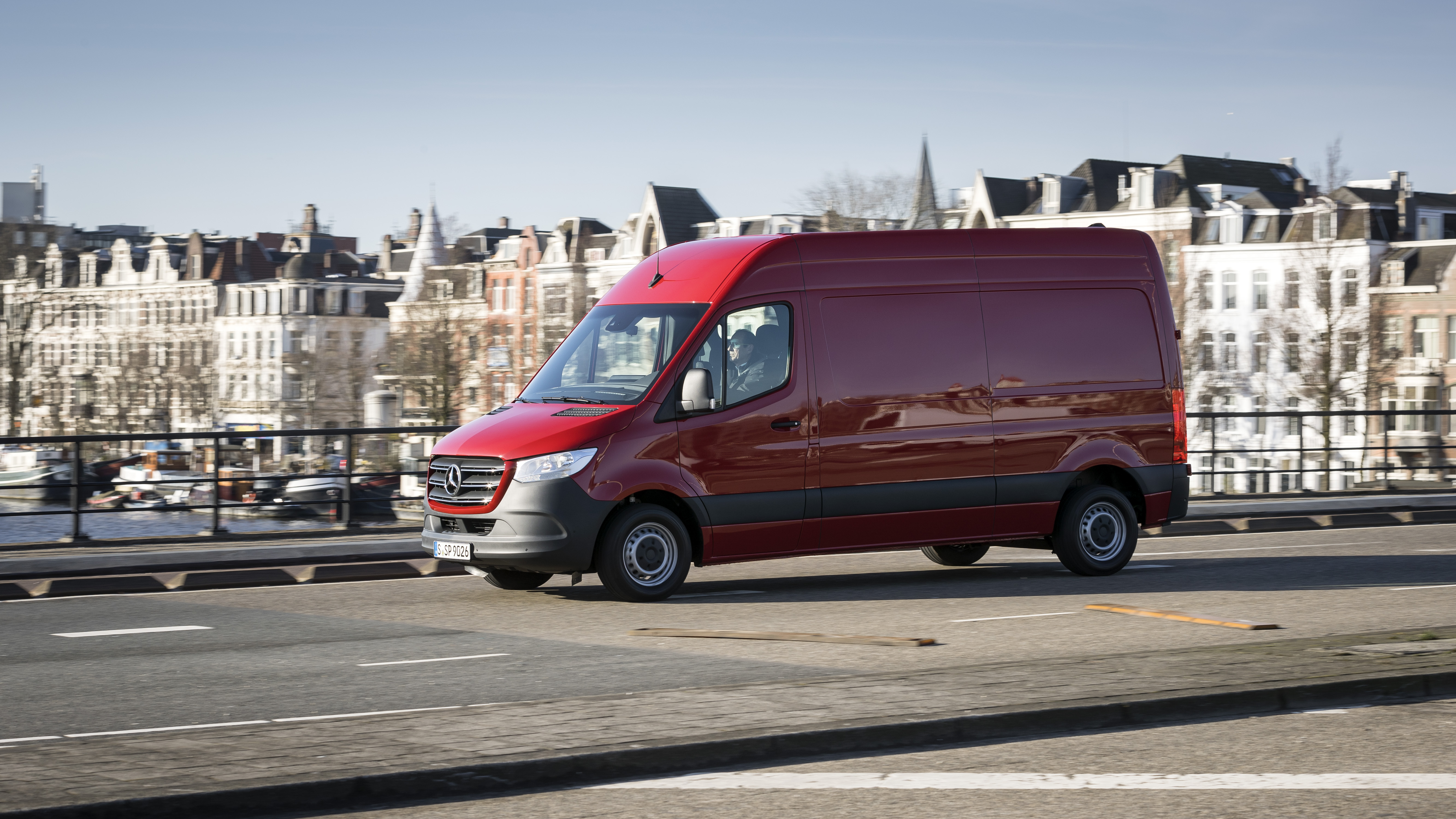 Ein roter Mercedes-Benz Sprinter fährt über eine Brücke vor historischer Stadtkulisse.