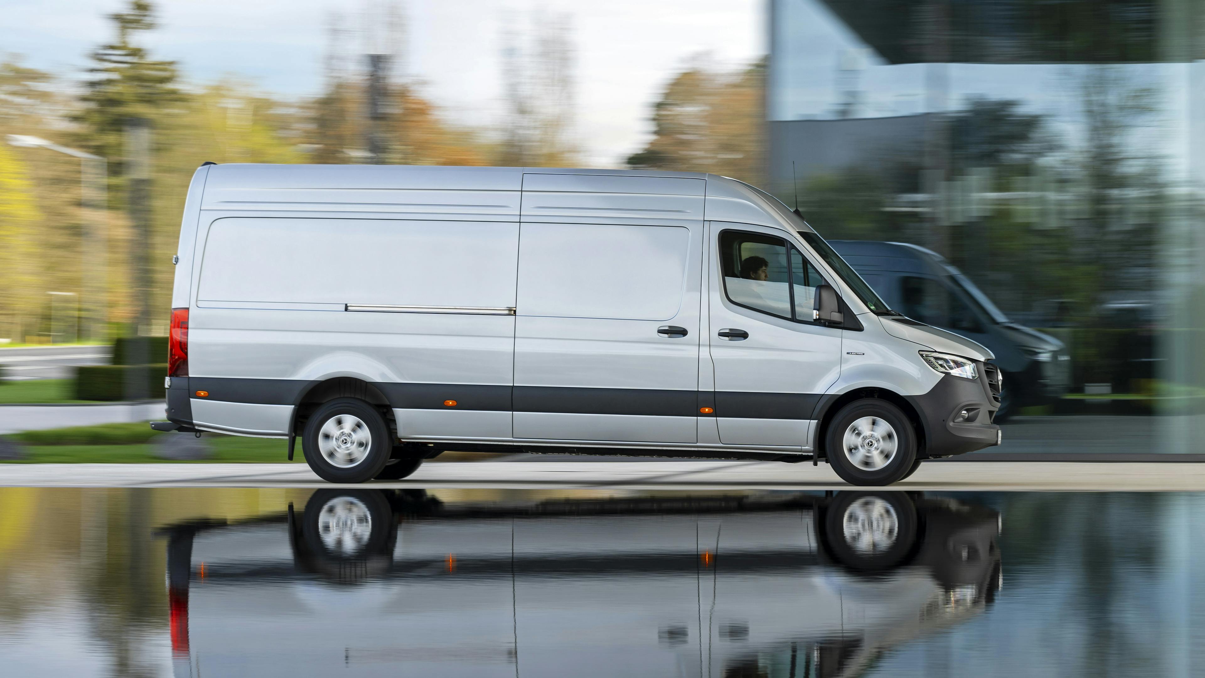 Ein silberner Mercedes-Benz Sprinter fährt an einem modernen Gebäude vorbei.