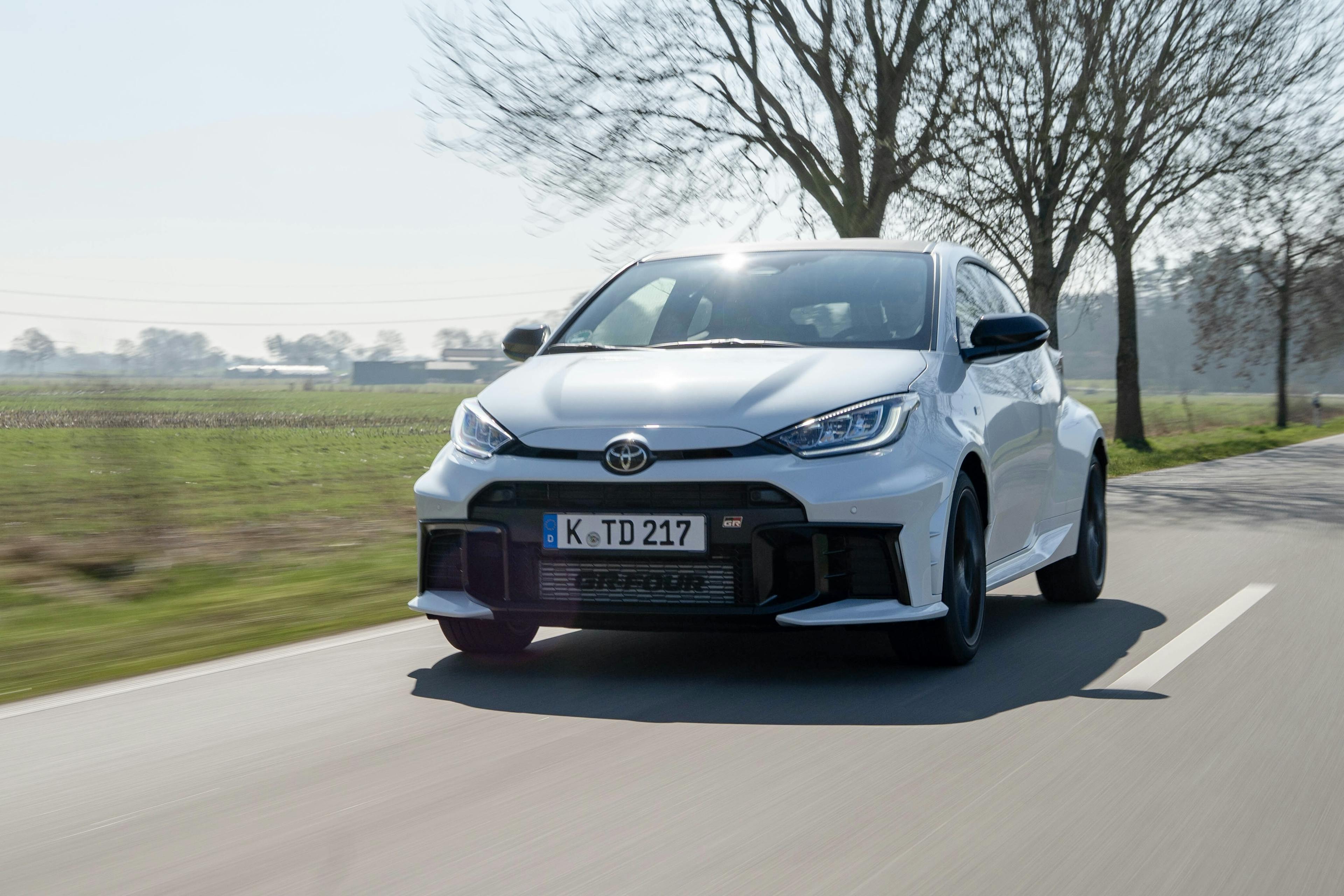 Ein weißer Toyota GR Yaris fahrend auf einer Landstraße vor laublosen Bäumen.