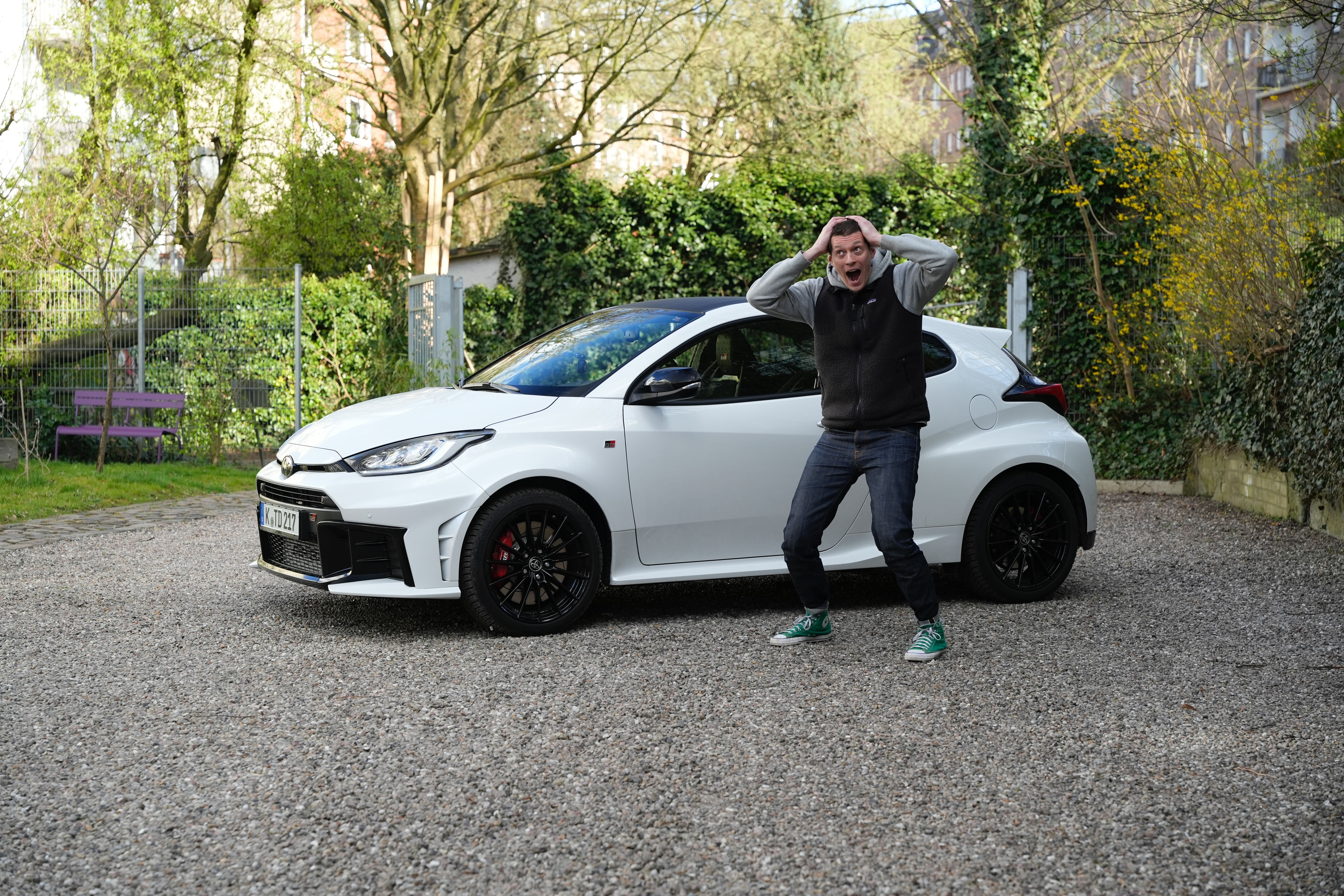 Ein weißer Toyota GR Yaris auf einem Kiesplatz stehend, mit einem verzückten Mann, der sich vor Begeisterung den Kopf hält.