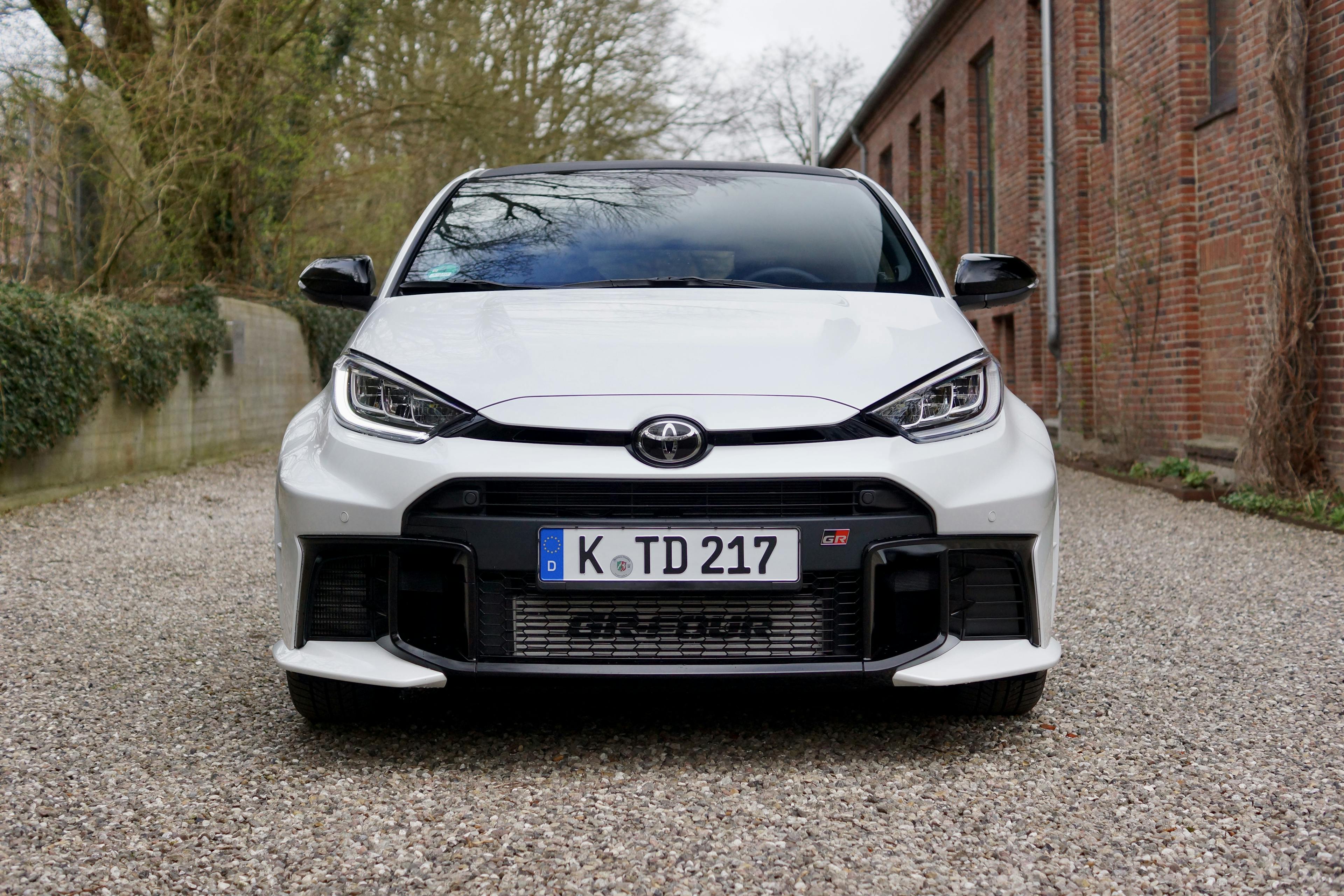 Ein weißer Toyota GR Yaris stehend auf einem Kiesplatz vor grüner Vegetation und einem Ziegelgebäude.