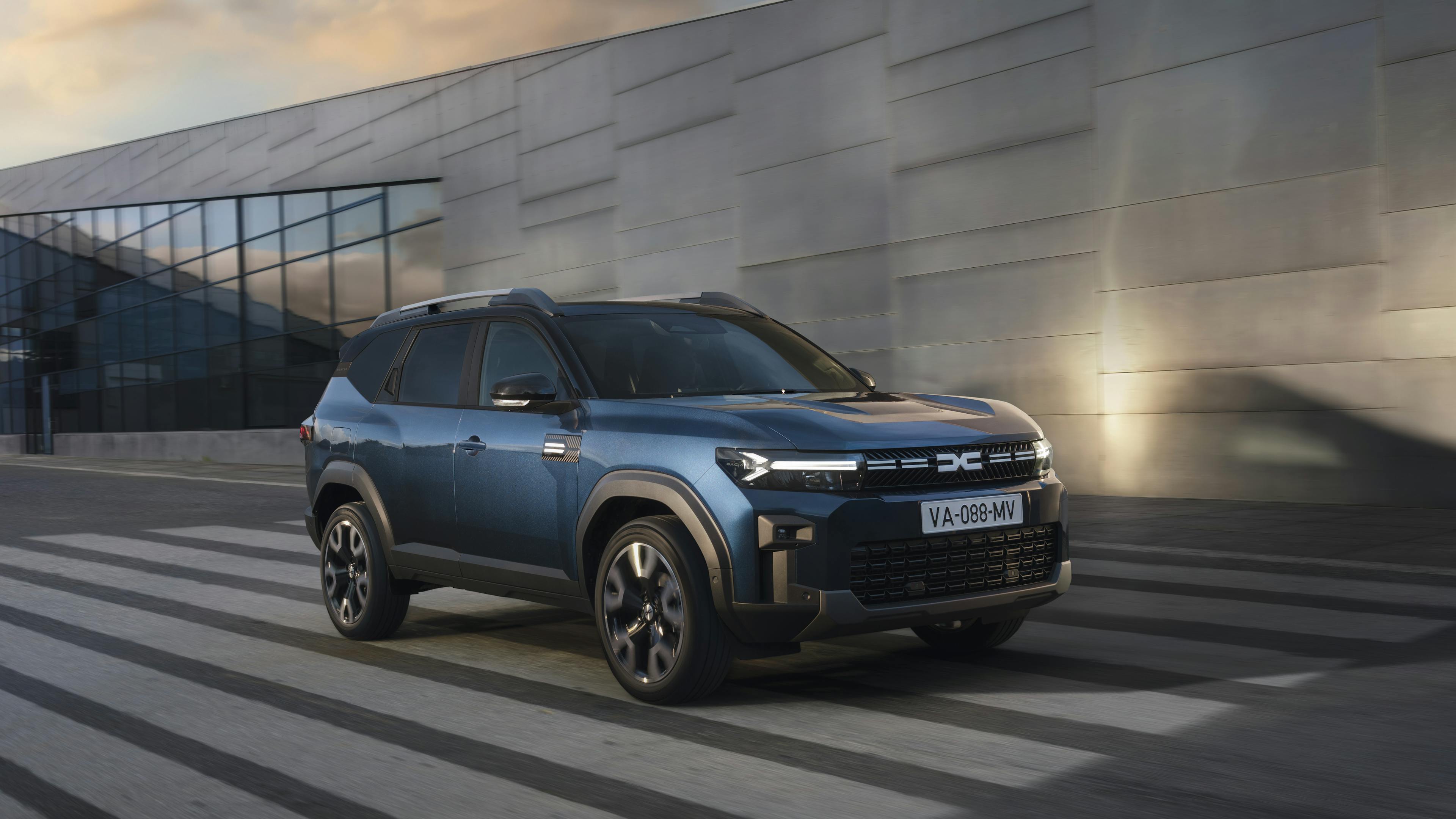 Ein blauer Dacia Bigster fährt an einer modernen Fassade vorbei.