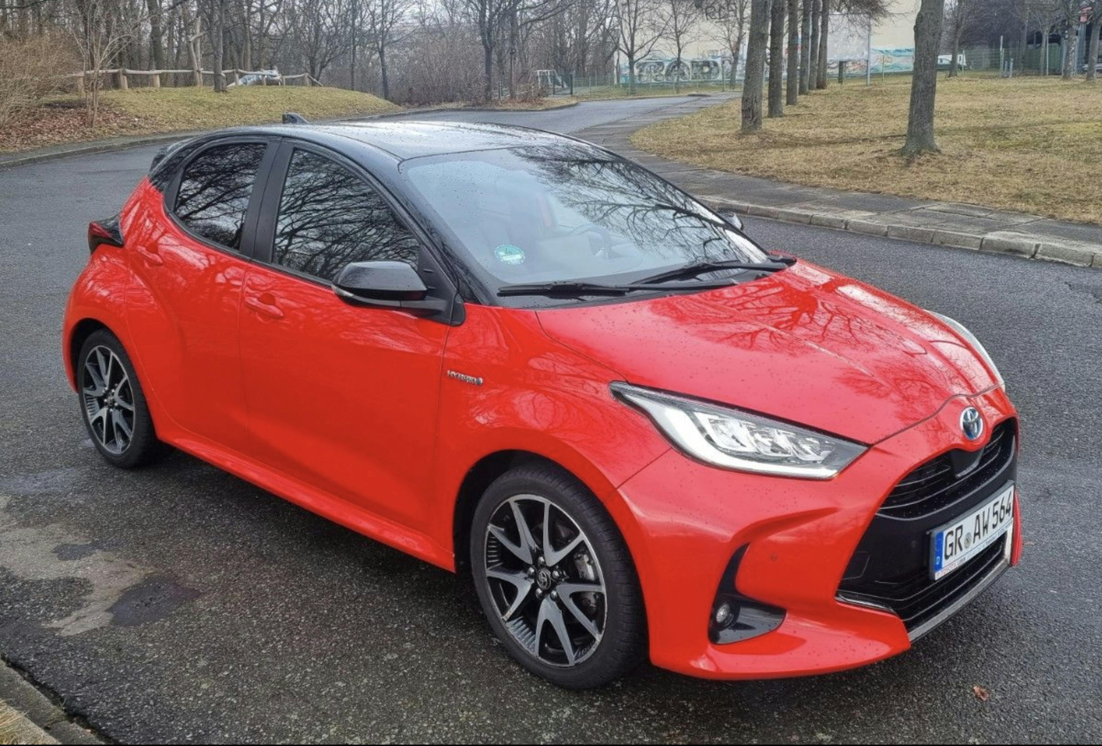 Roter Toyota Yaris auf einer Asphaltfläche stehend, mit winterlicher Vegetation im Hintergrund.