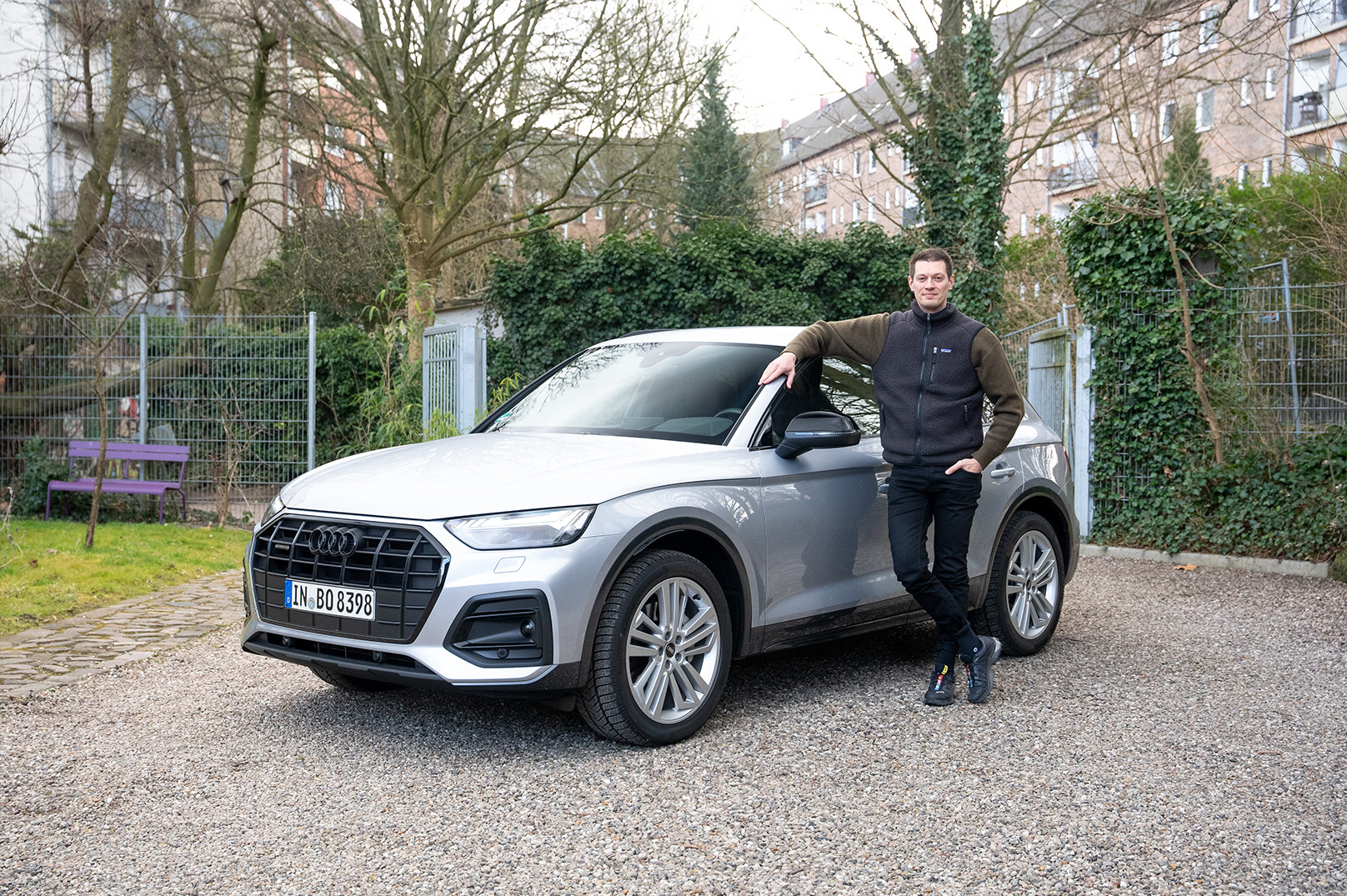 Die Frontansicht eines silbernen Audi Q5 FY 50 TDI quattro auf einem Kiesplatz mit einem Mann, der sich keck an die Fahrertür lehnt.