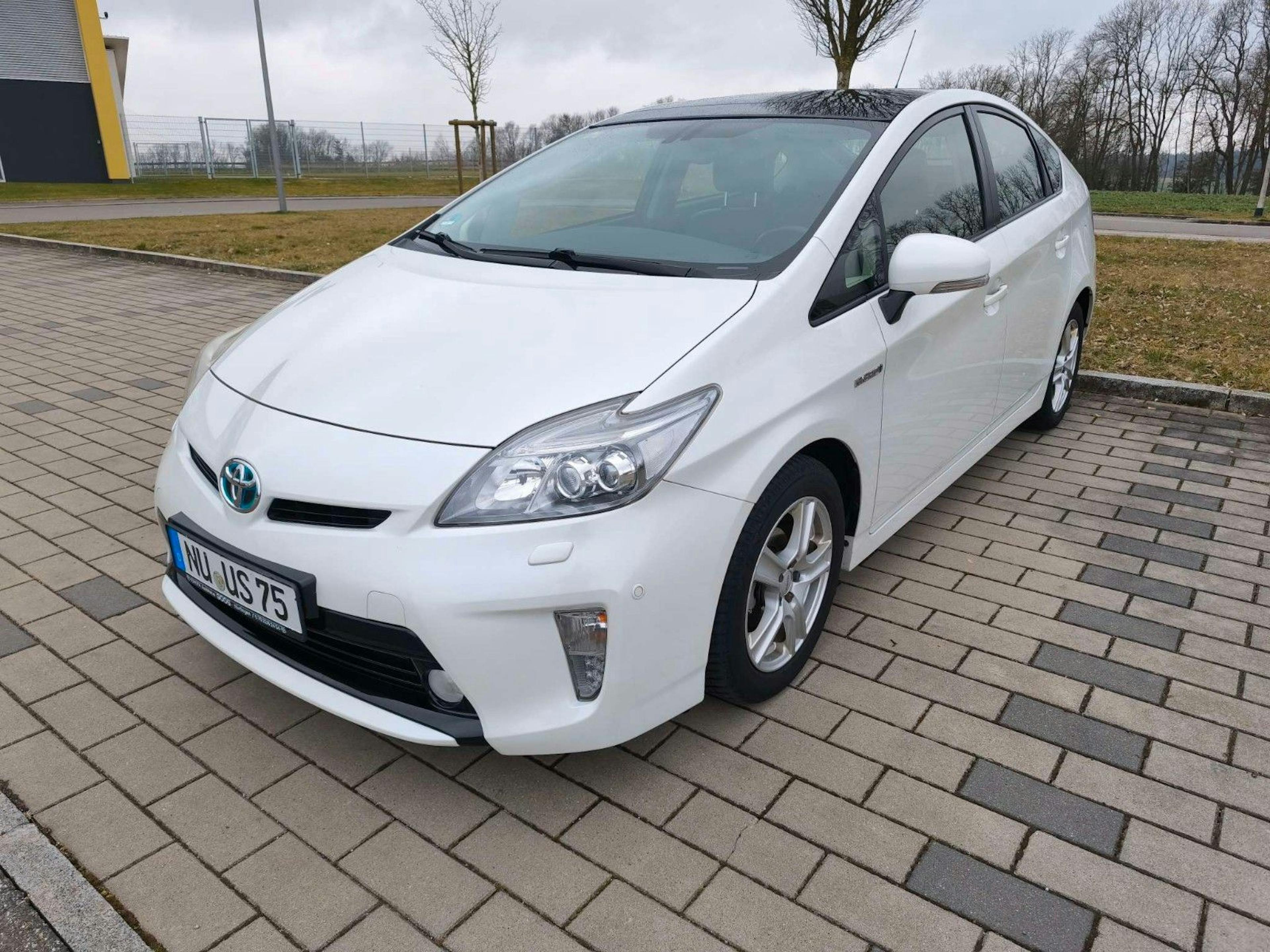 Ein weißer Toyota Prius steht auf einem gepflasterten Parkplatz mit kahlen Bäumen.