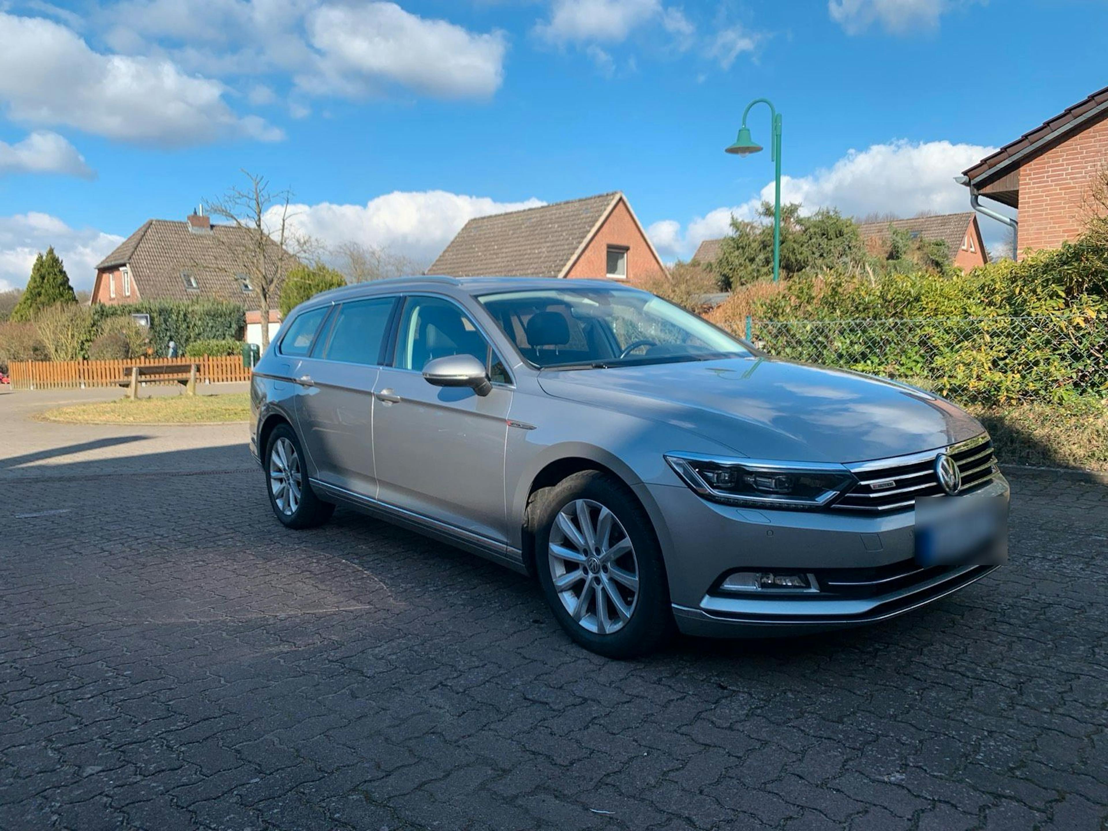 Ein silberner Volkswagen Passat Variant steht auf einem gepflasterten Parkplatz in einem Wohngebiet mit Einfamilienhäusern und Hecken im Hintergrund.