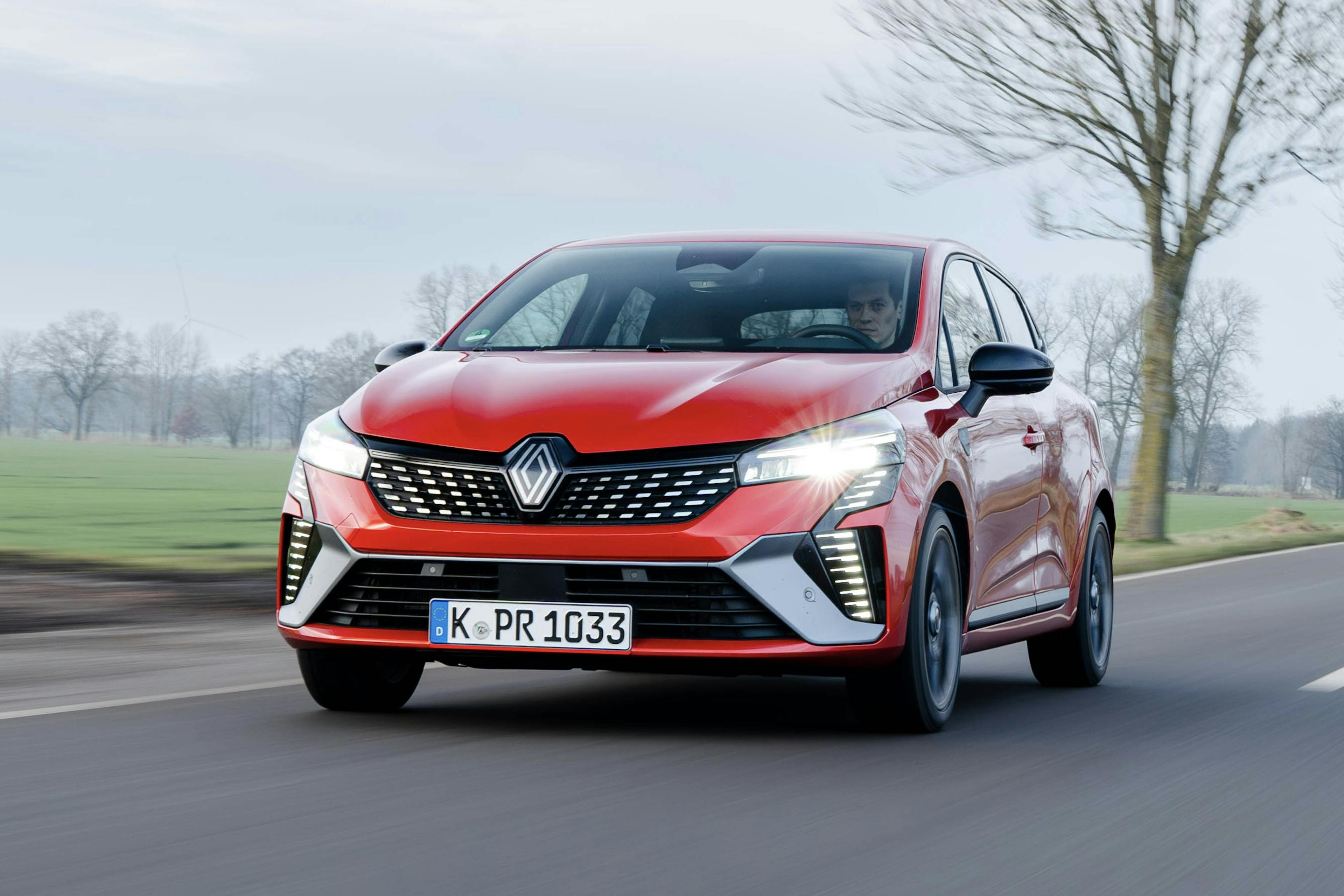 Frontansicht eines roten Renault Clio auf einer Landstraße, fahrend mit laublosen Bäumen im Hintergrund.