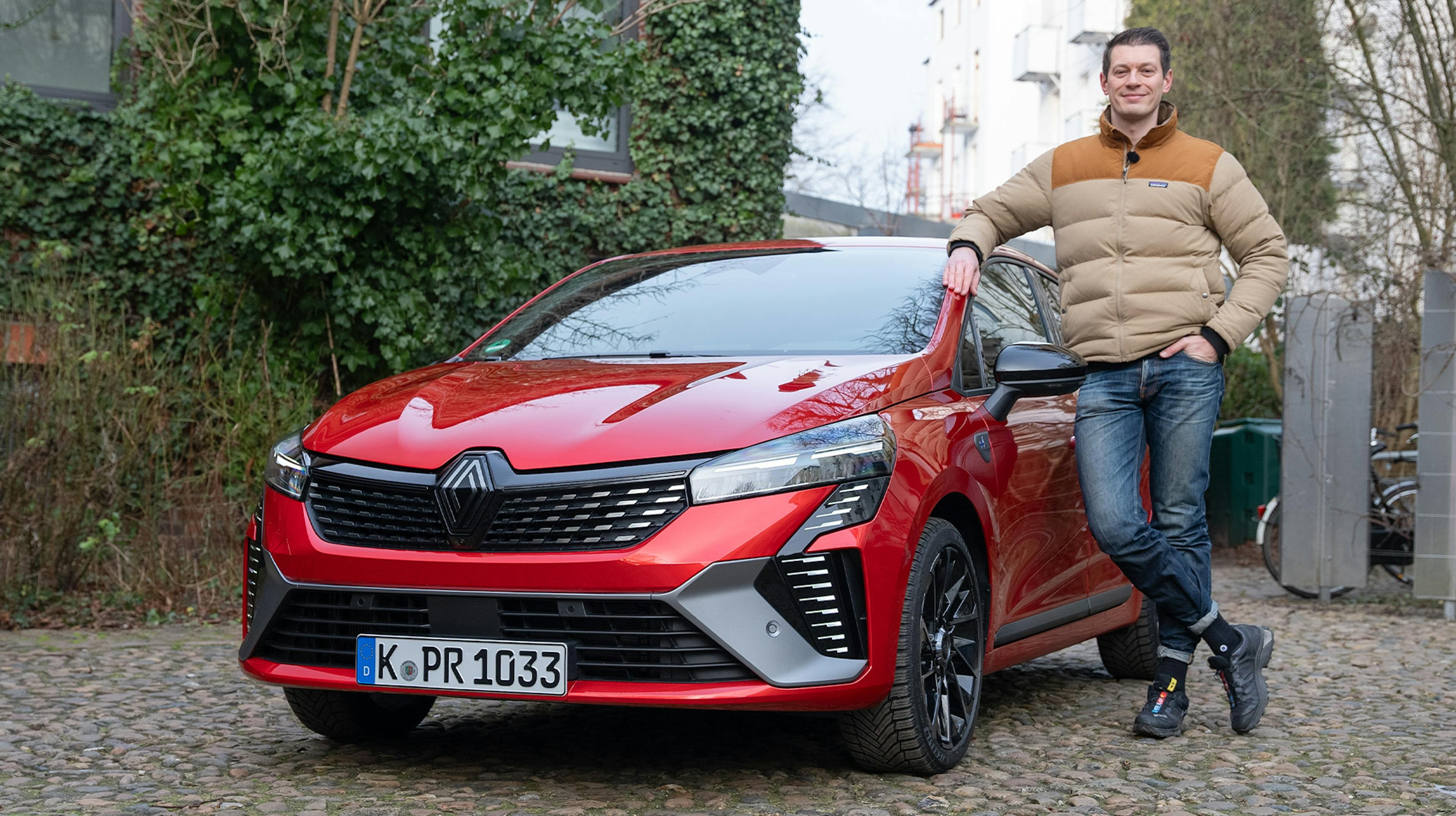 Ein roter Renault Clio auf einem Kopfsteinpflasterplatz vor einem begrünten Gebäude, mit einem Mann ans Auto gelehnt.