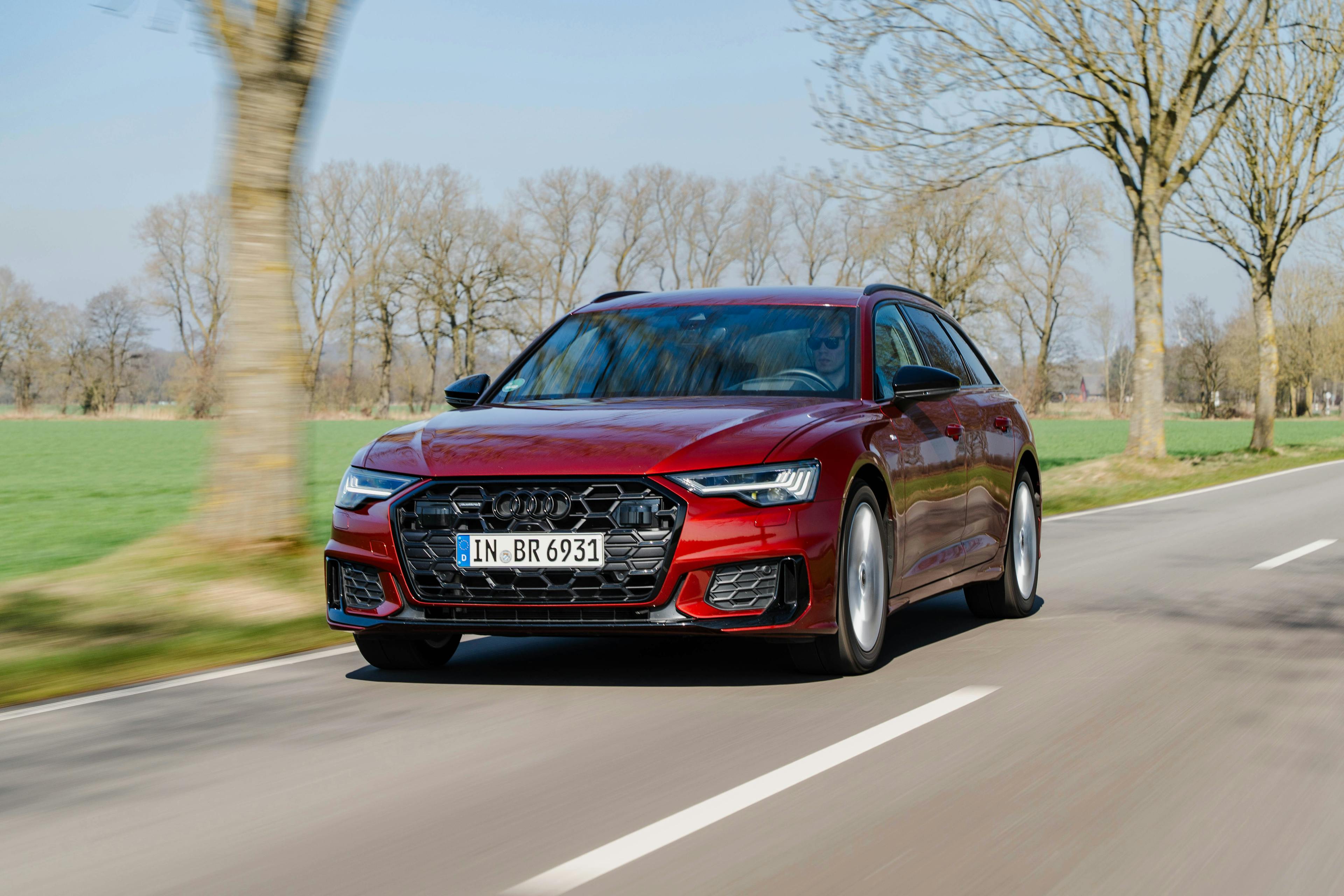 Roter Audi A6 Avant C8 fahrend auf einer Landstraße vor laublosen Bäumen.