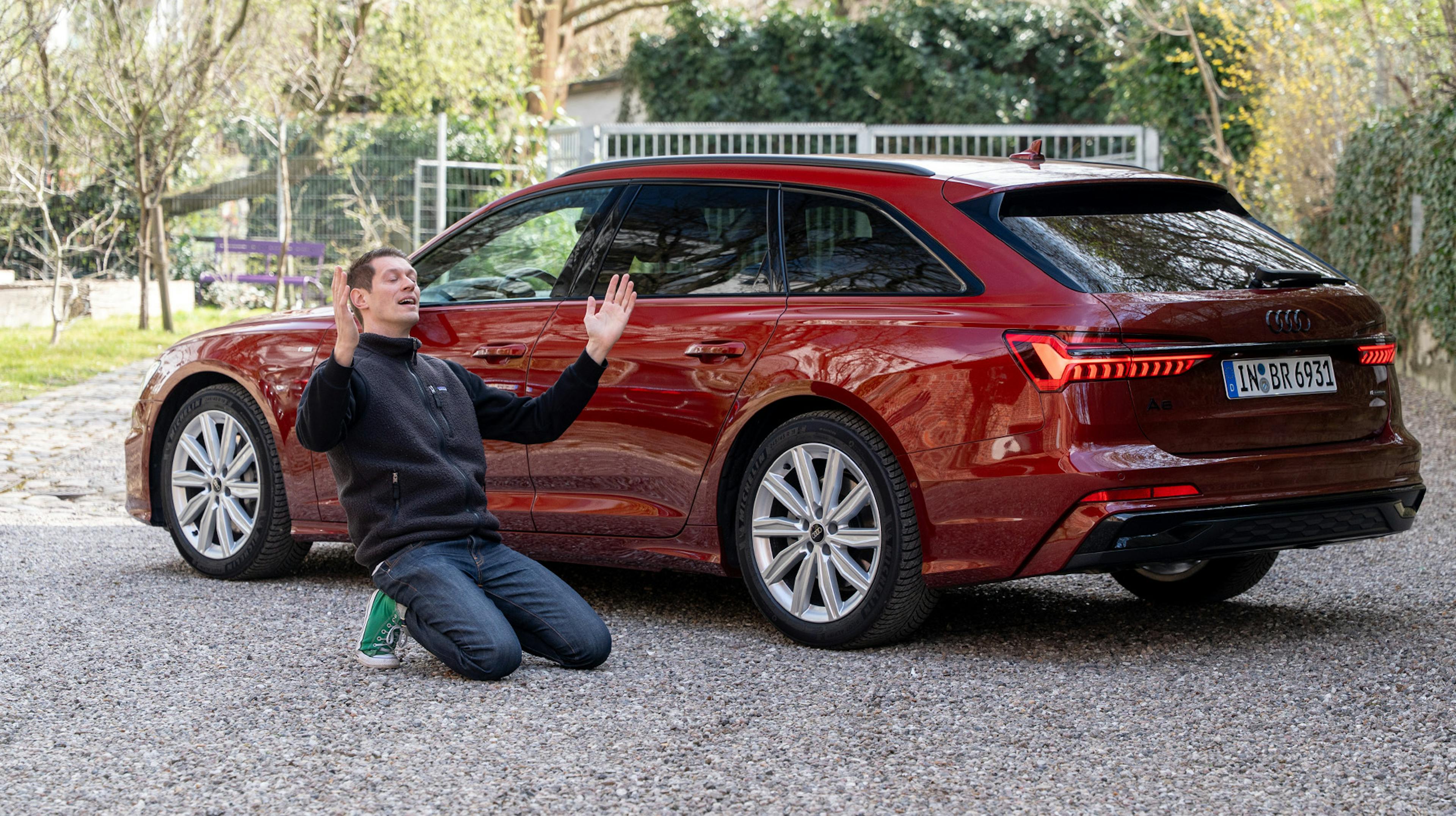 Die Frontansicht eines roten Audi A6 Avant 50 TDI quattro auf einem Kiesplatz mit einem Mann, der begeistert vor dem Auto kniet.