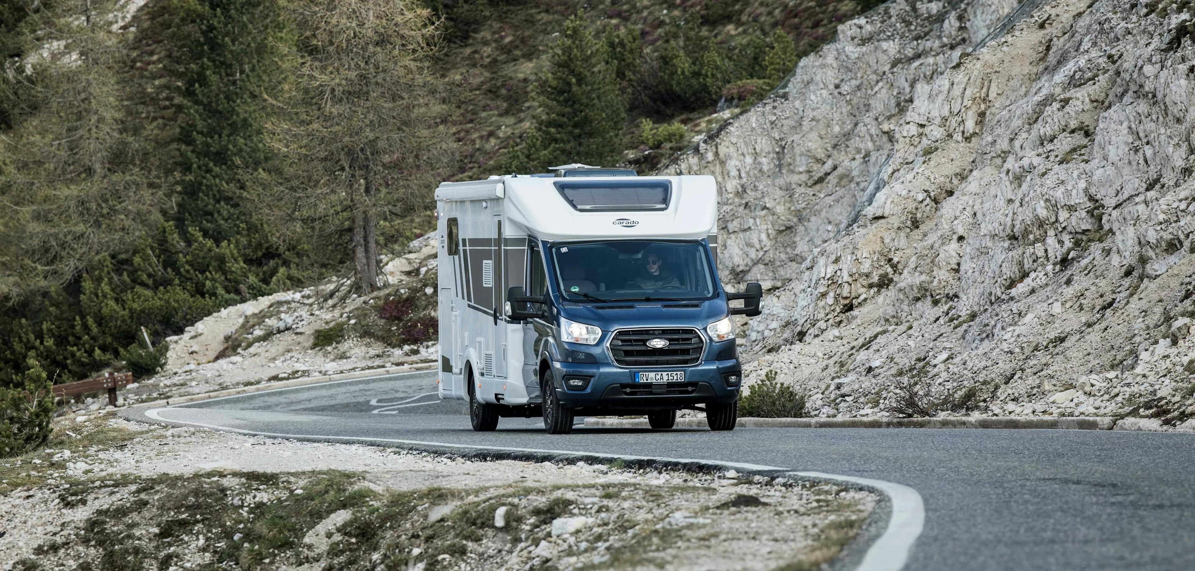 Ein weiß-blaues Carado-Wohnmobil fährt auf einer kurvigen Bergstraße mit felsiger Landschaft und Nadelbäumen.