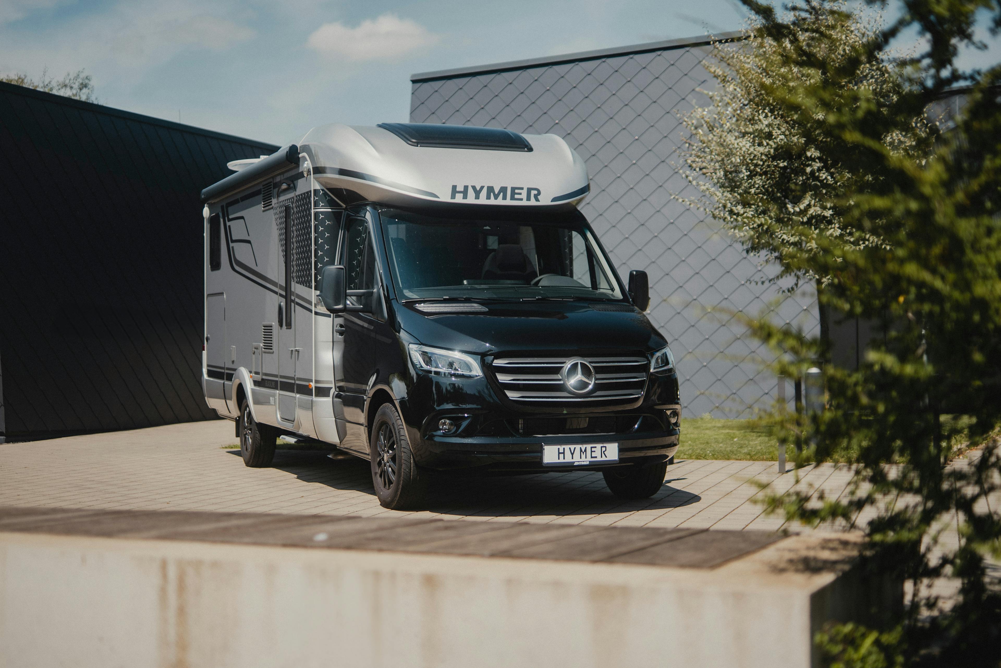 Ein modernes Hymer-Wohnmobil auf Mercedes-Basis steht auf einem gepflasterten Platz vor einem modernen Gebäude.