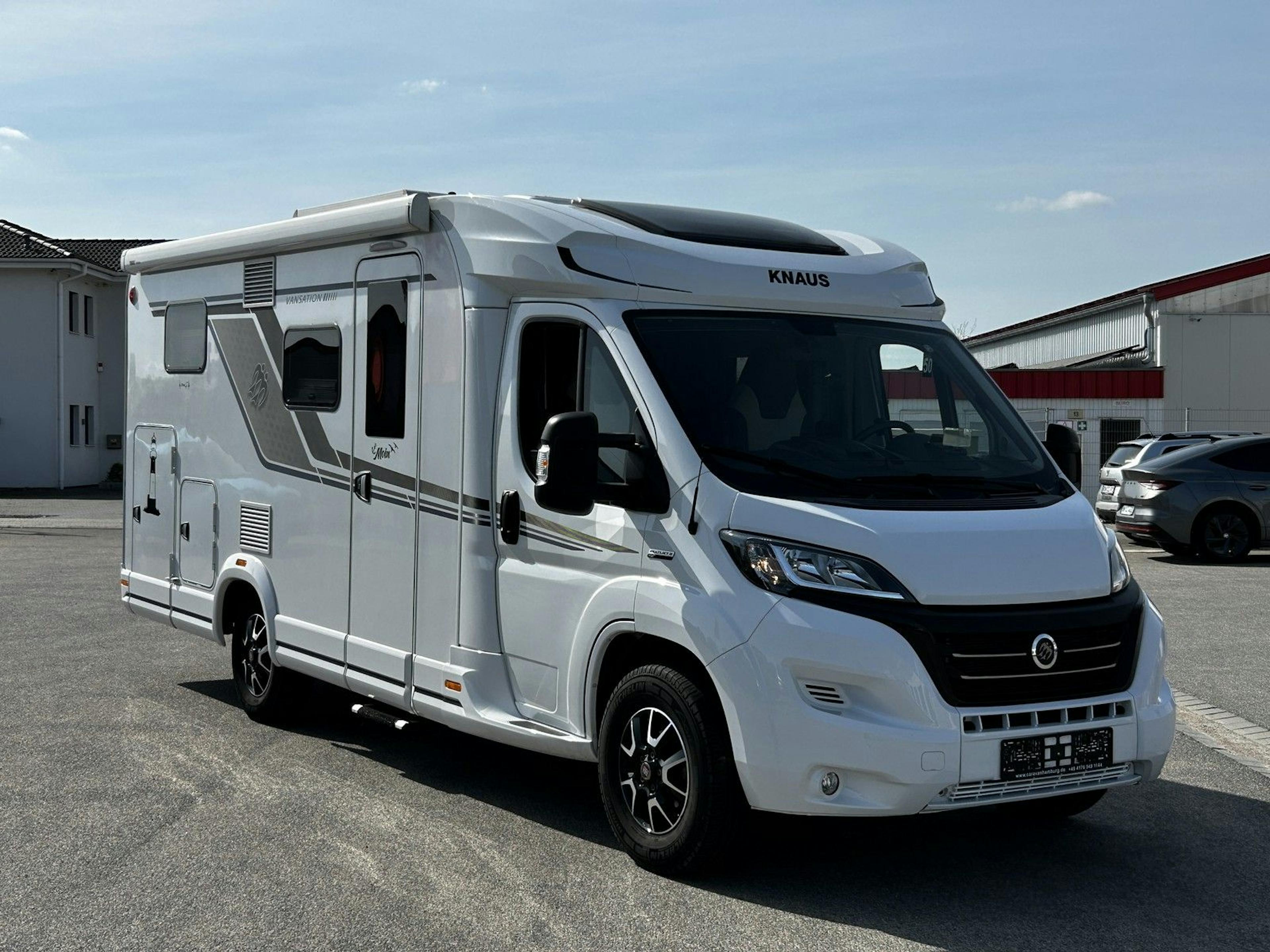 Modernes, teilintegriertes Wohnmobil von Knaus auf Fiat-Ducato-Chassis, geparkt auf einem befestigten Platz vor einer Halle.