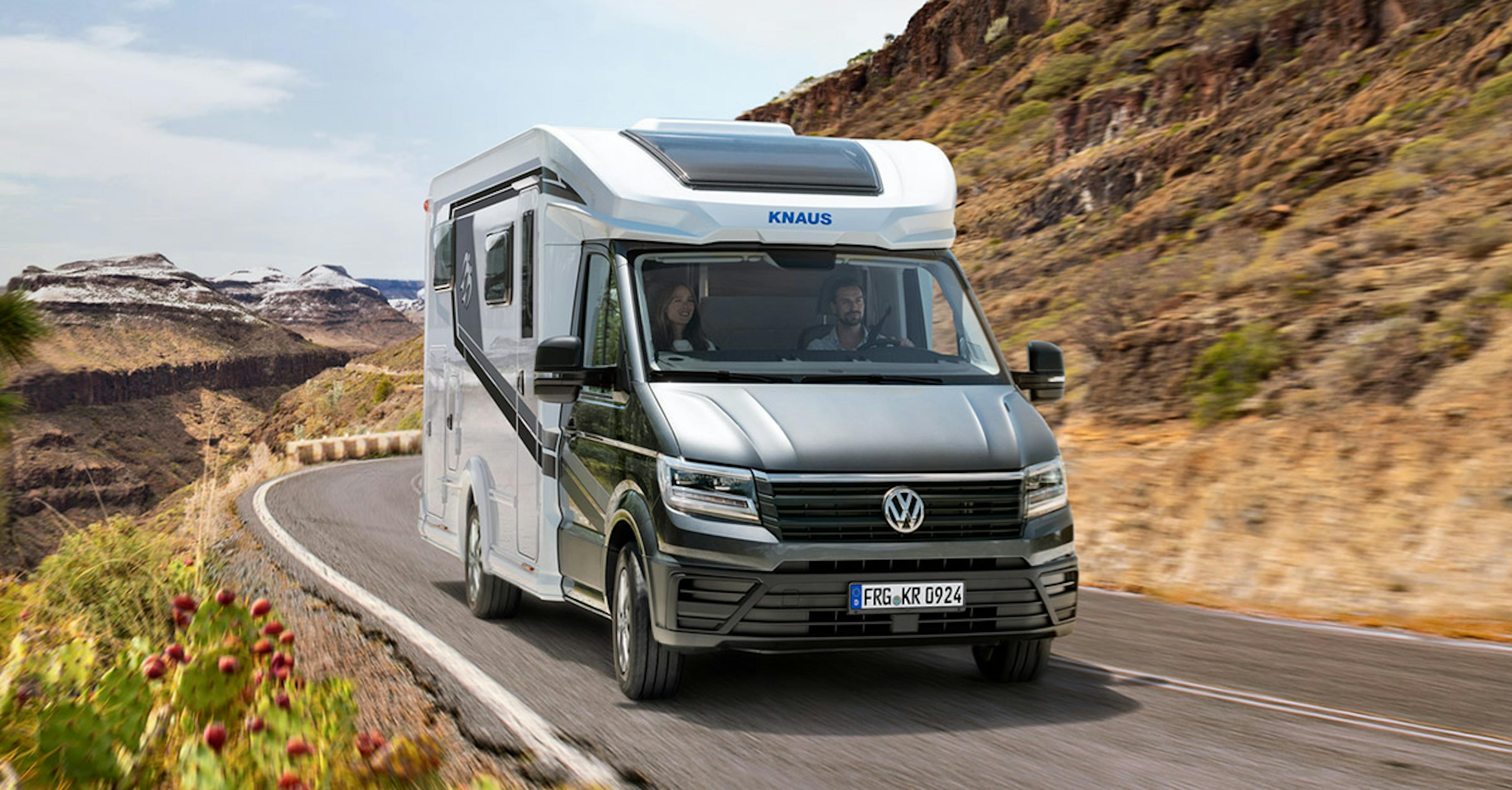 Ein Knaus-Wohnmobil mit VW-Front fährt durch eine kurvige Straße in einer trockenen, bergigen Landschaft.