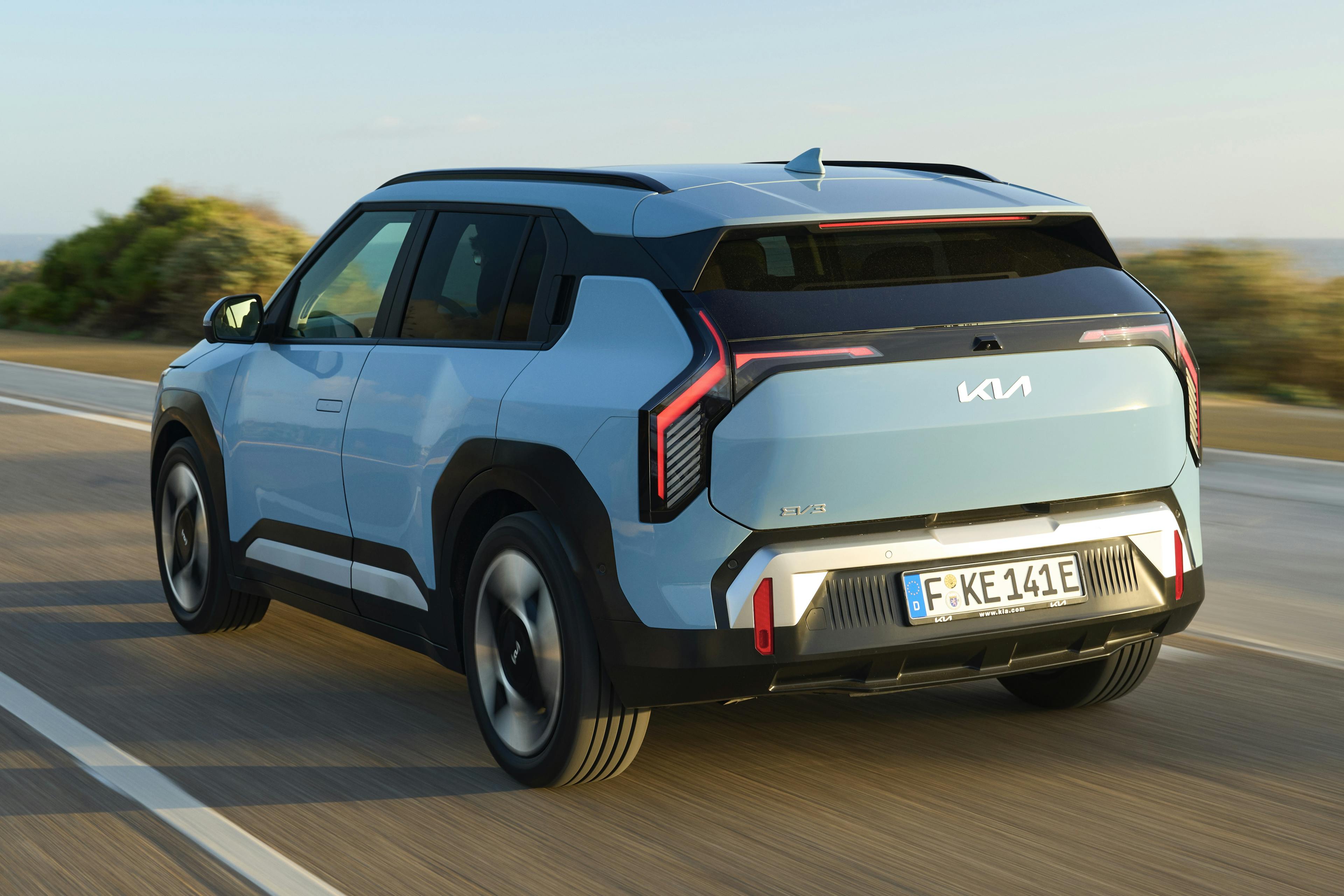 Blick auf das Heck eines fahrenden hellblauen Kia EV3 auf einer Landstraße.