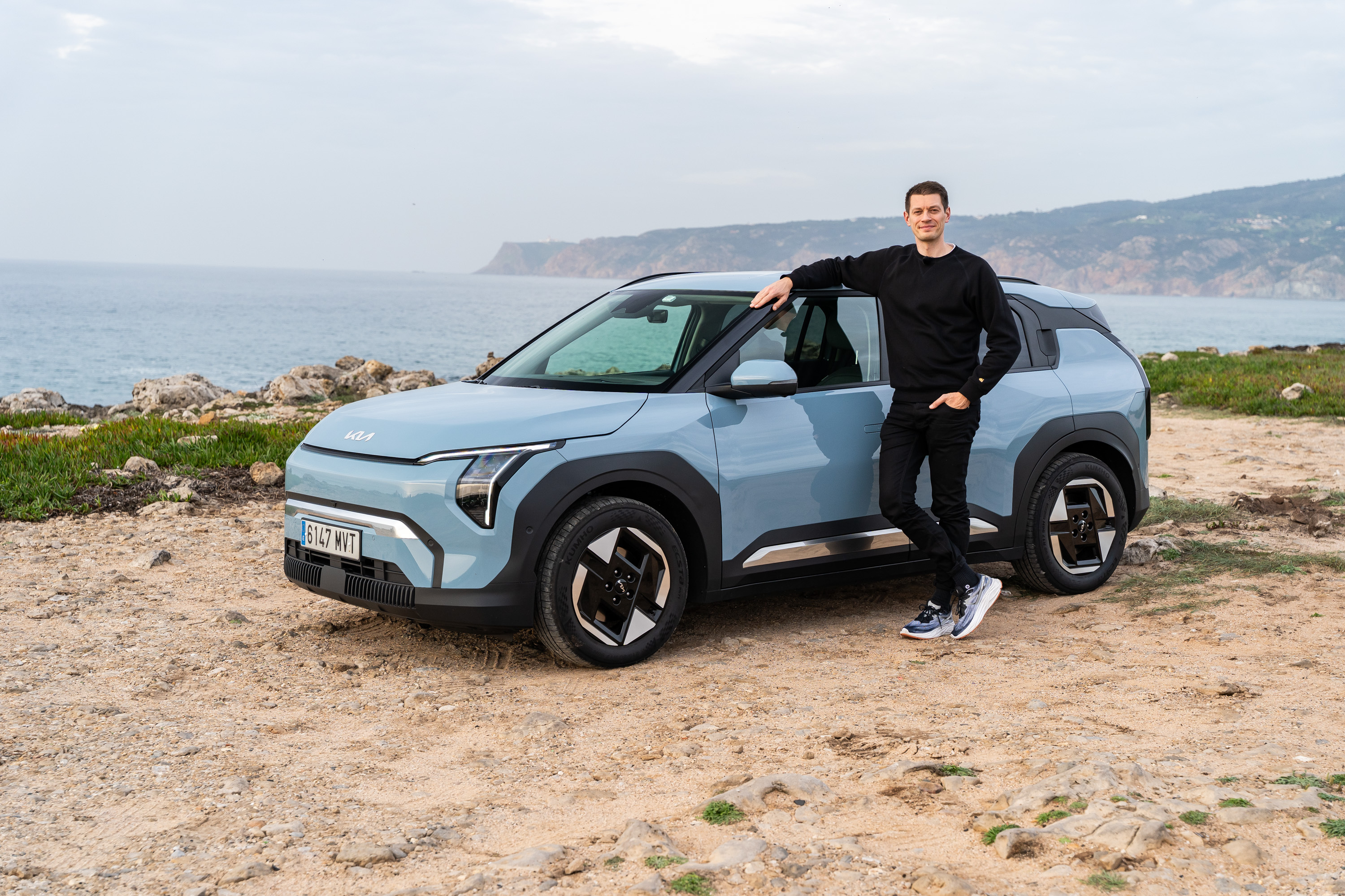 Hellblauer Kia EV3 an einem Strand stehend, mit einem ans Auto gelehnten Mann, der zufrieden aussieht.