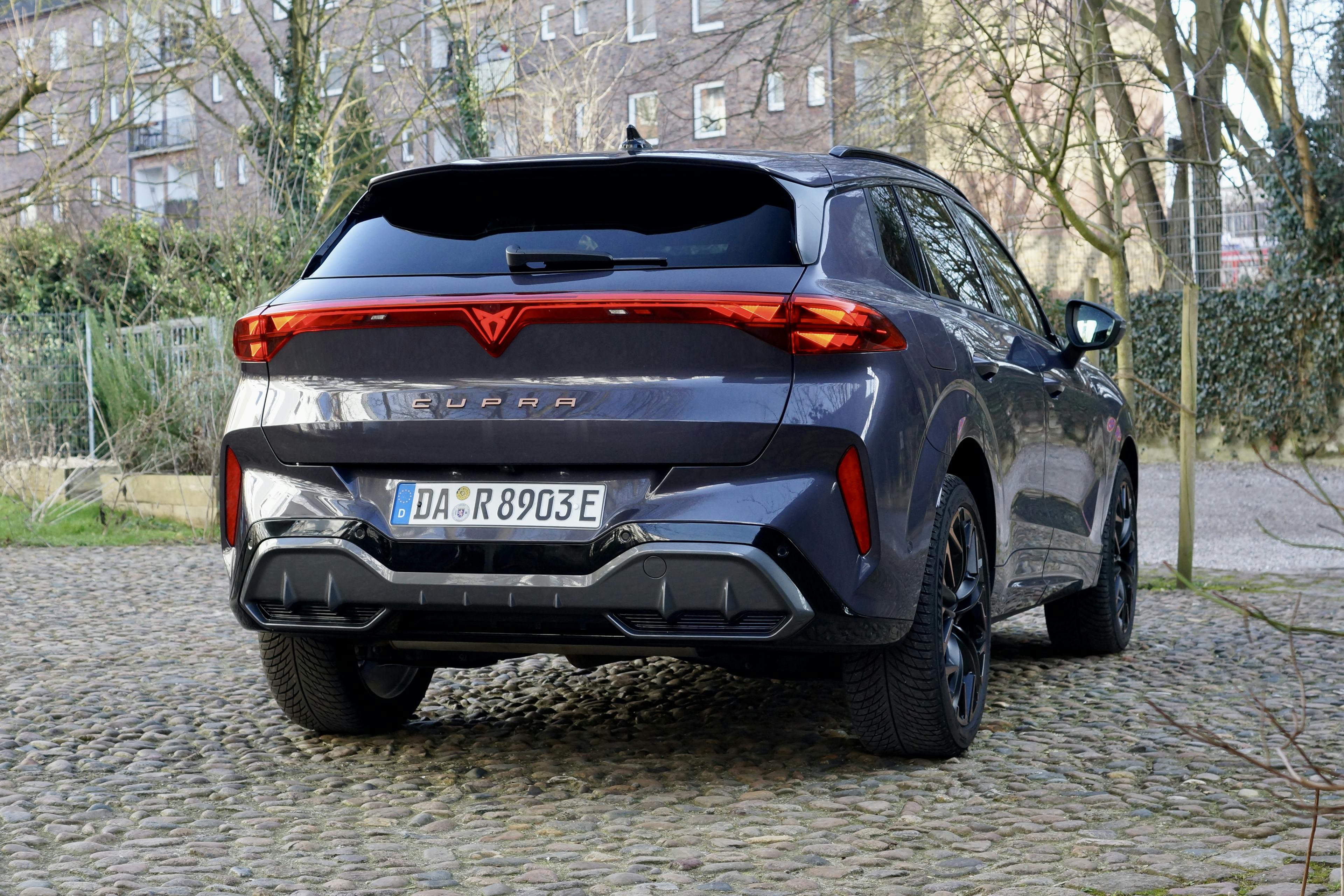 Heckansicht eines stehenden lilafarbenen Cupra Terramar auf einem Kopfsteinpflasterplatz vor einem Ziegelgebäude.
