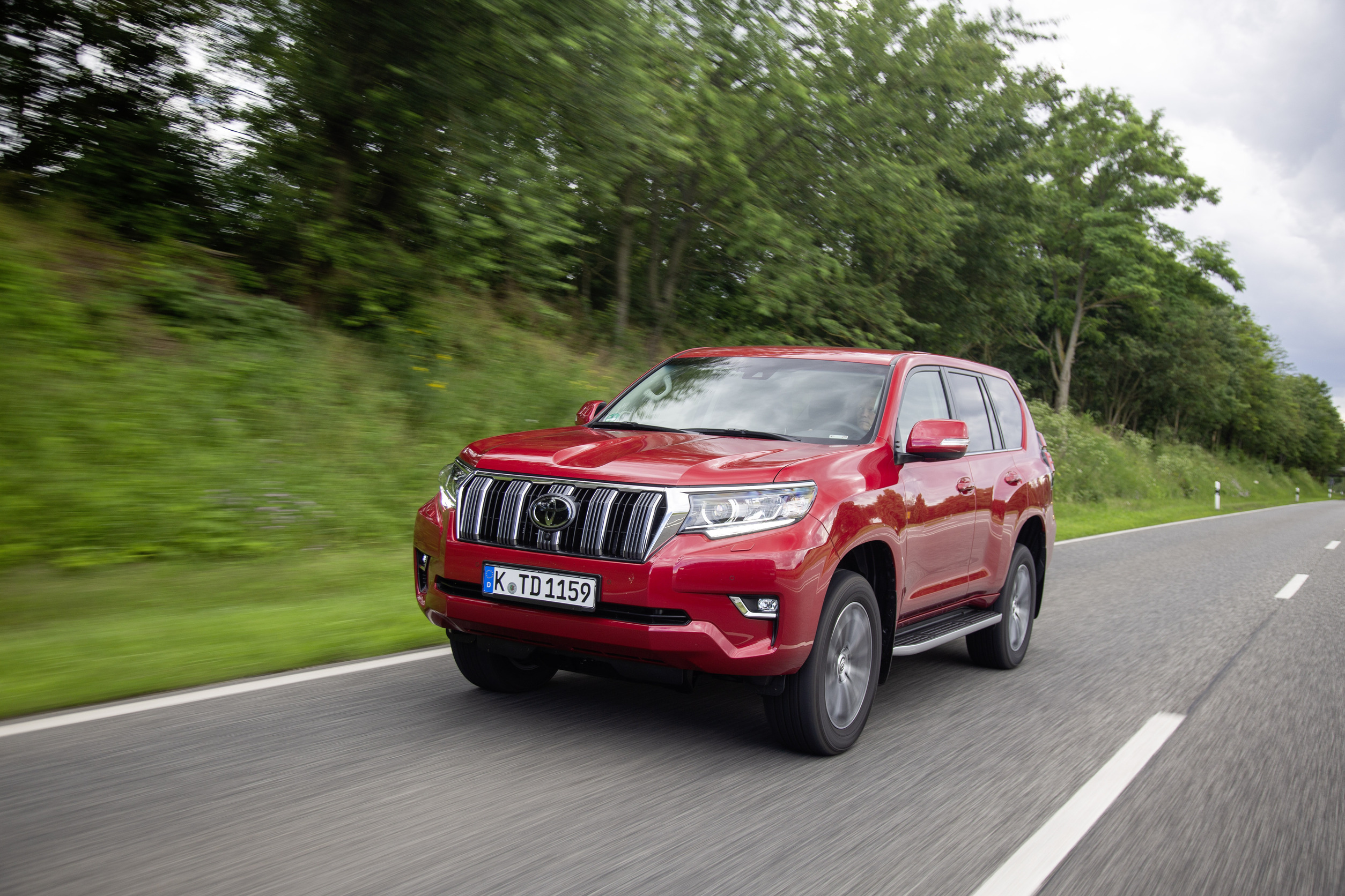 Ein roter Toyota Land Cruiser fährt auf einer Straße mit Bäumen im Hintergrund.
