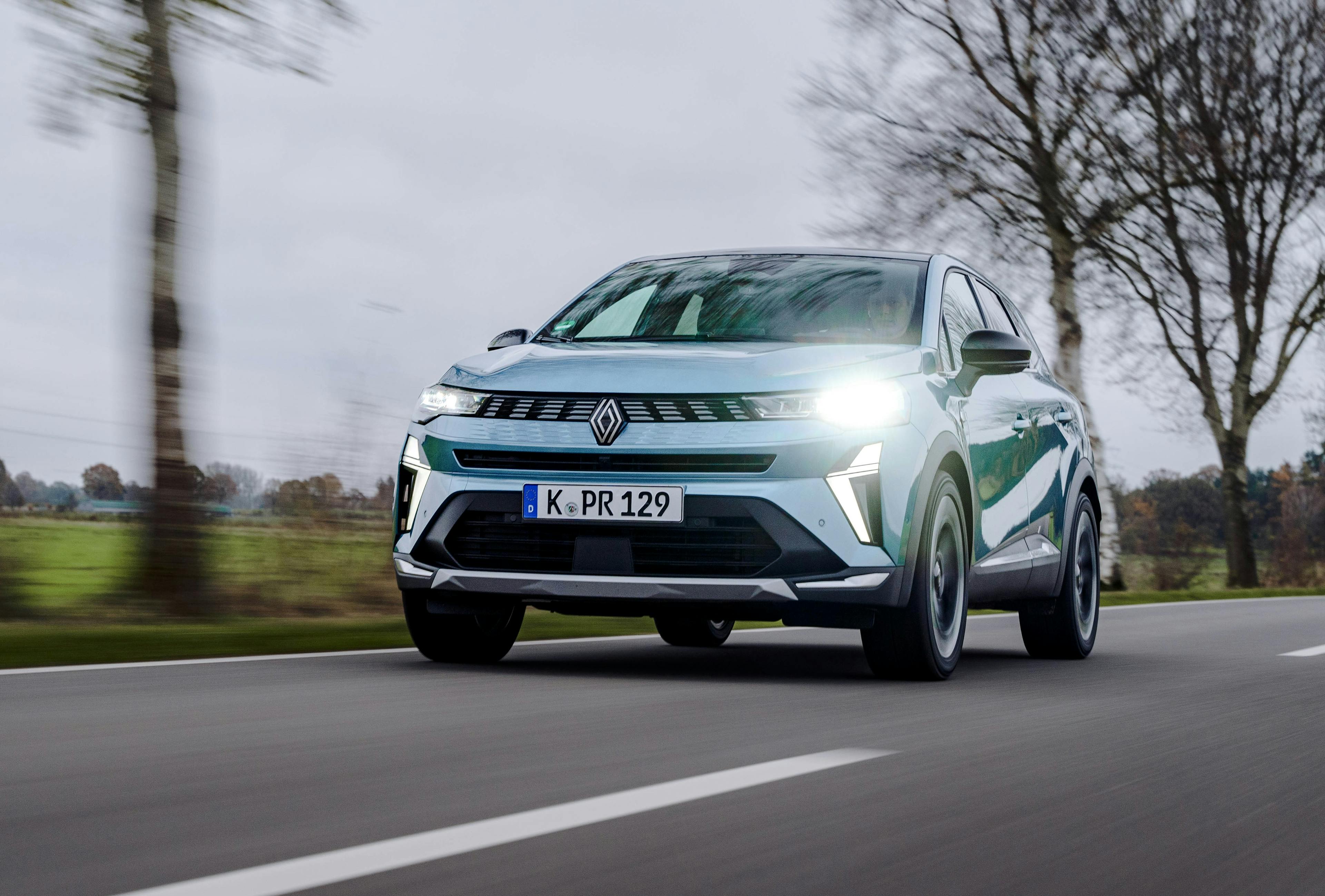 Frontansicht eines blauen Renault Symbioz auf einer Landstraße, fahrend mit laublosen Bäumen im Hintergrund