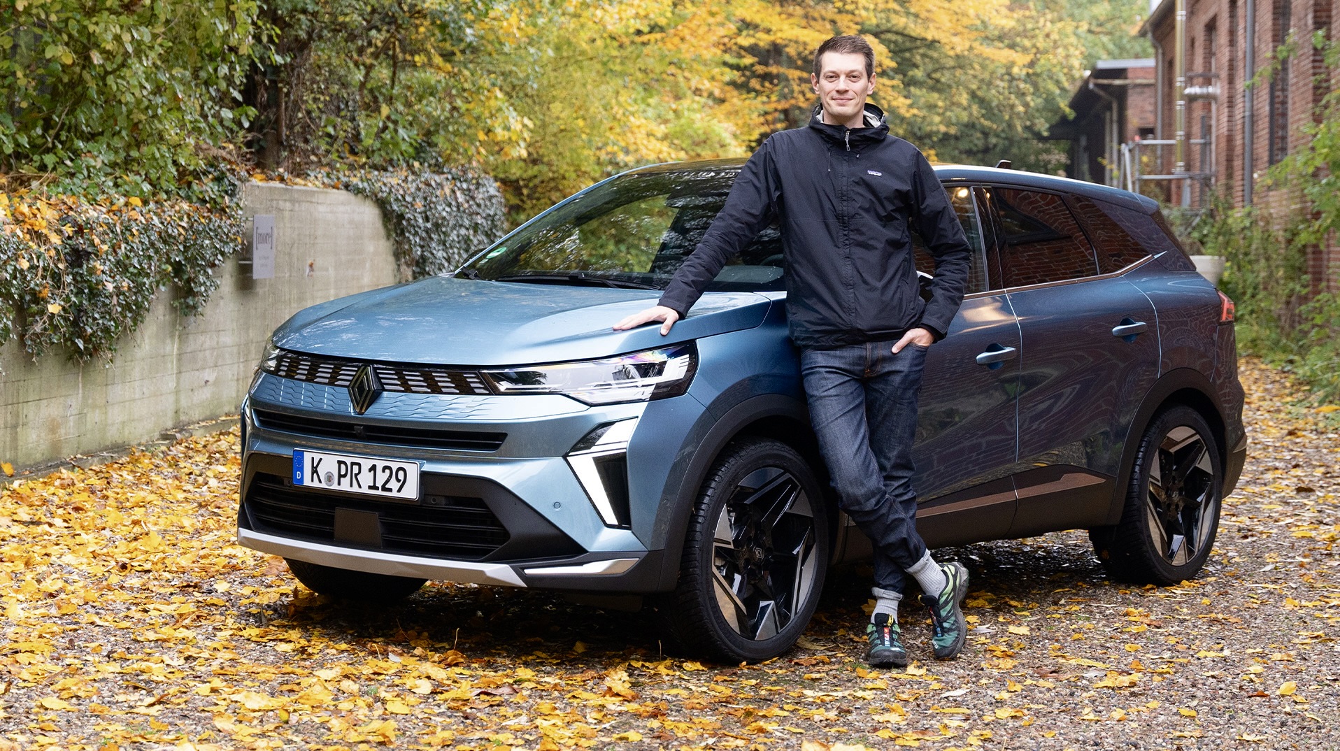 Ein blauer Renault Symbioz auf einem herbstlichen Kopfsteinpflasterplatz, mit einem Mann ans Auto gelehnt.