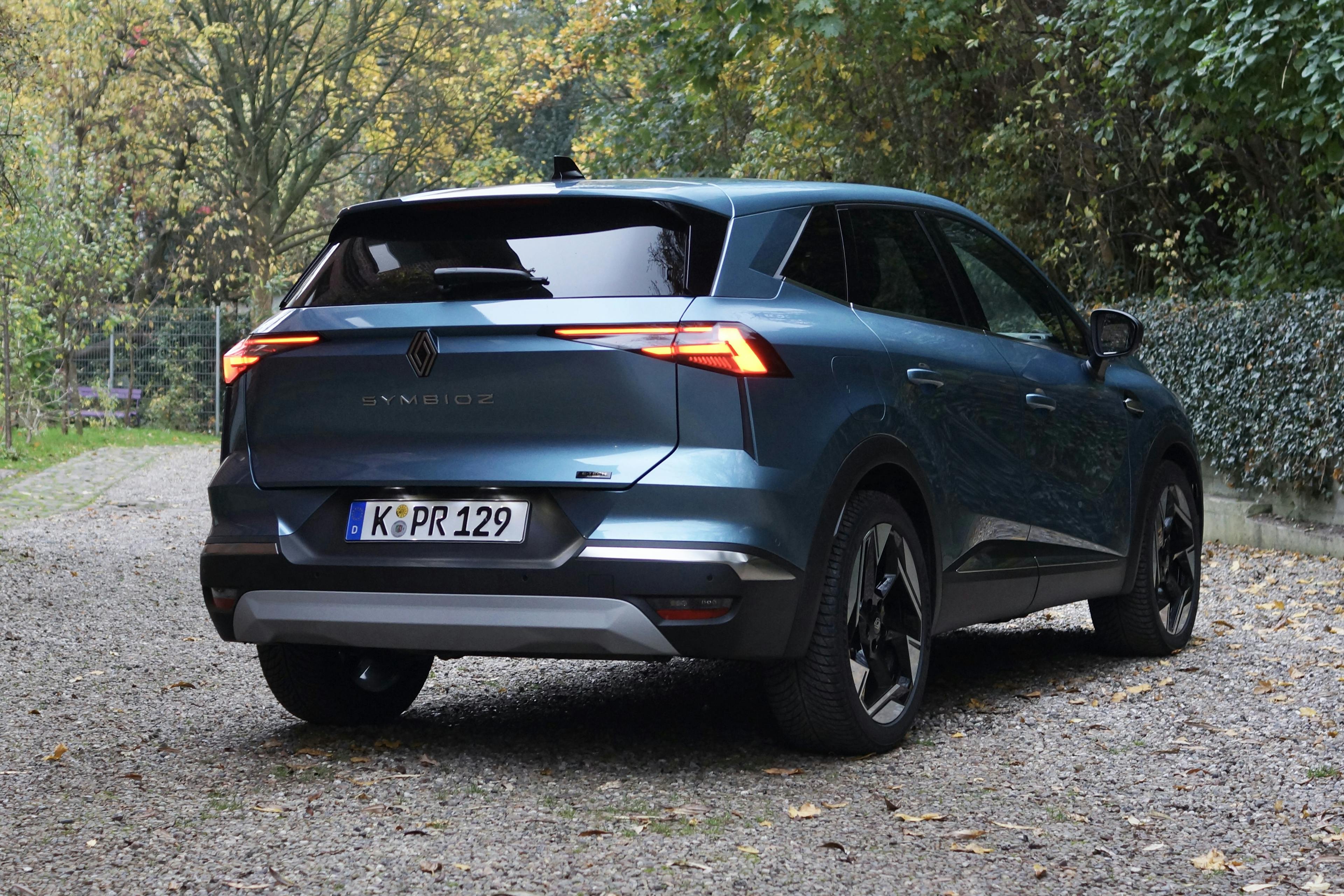 Heckansicht eines blauen Renault Symbioz auf einem Kiesplatz vor herbstlicher Bepflanzung.