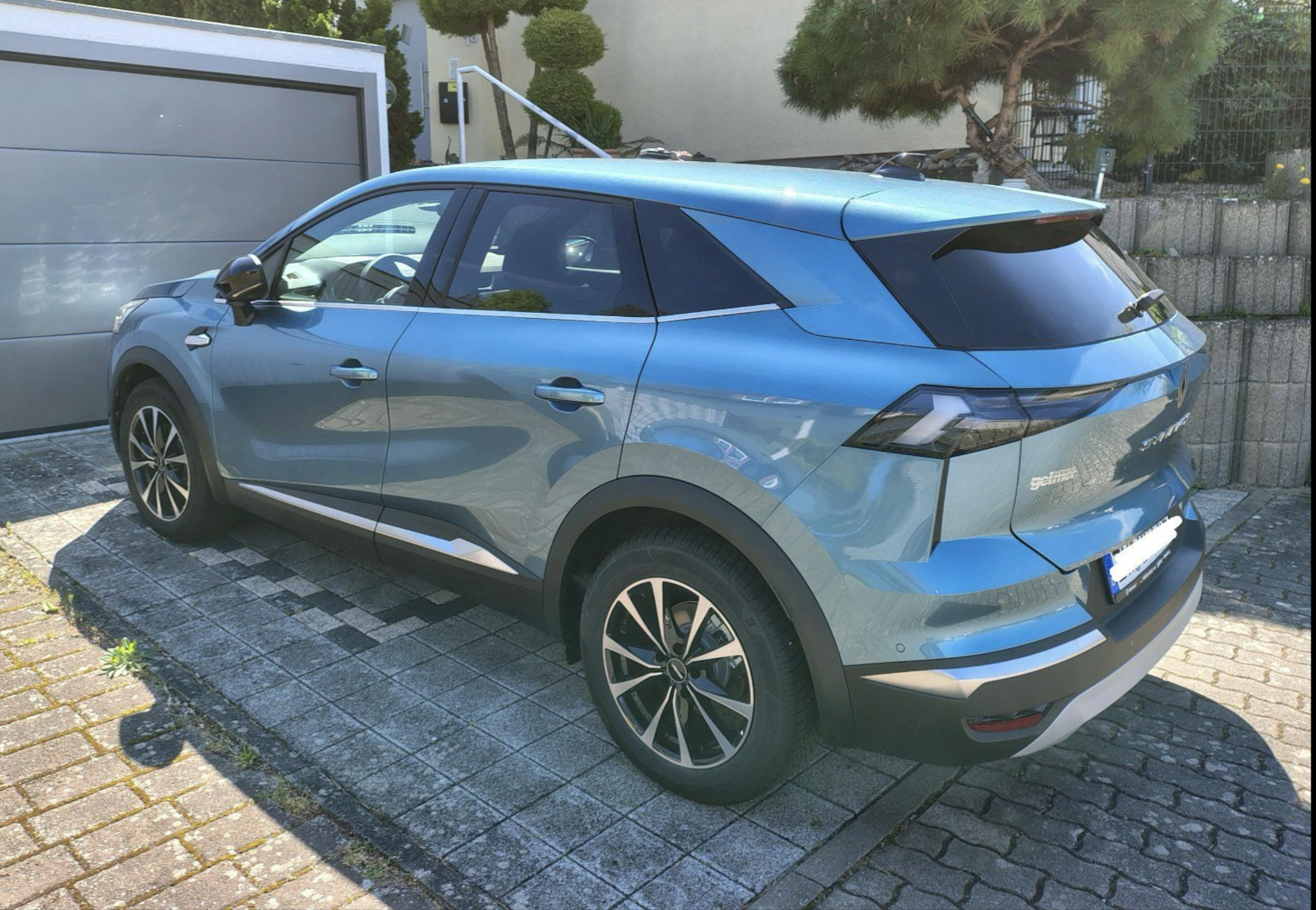 Blauer Renault Symbioz, stehend auf einer gepflasterten Fläche vor einer Garage.