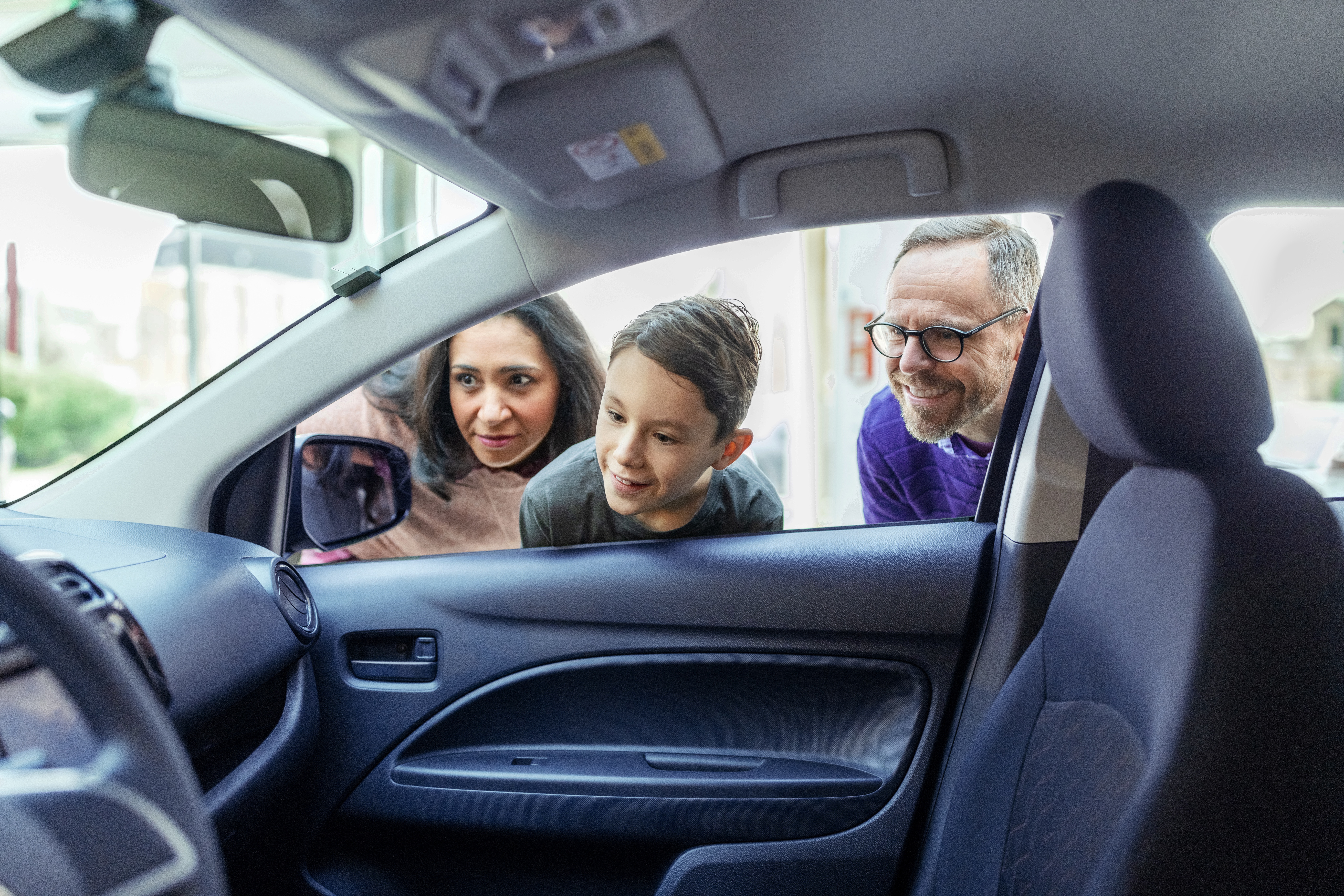 Eine Frau, ein Mann und ein Kind schauen gemeinsam von außen durch die geöffnete Autoscheibe in den Innenraum eines Fahrzeugs.