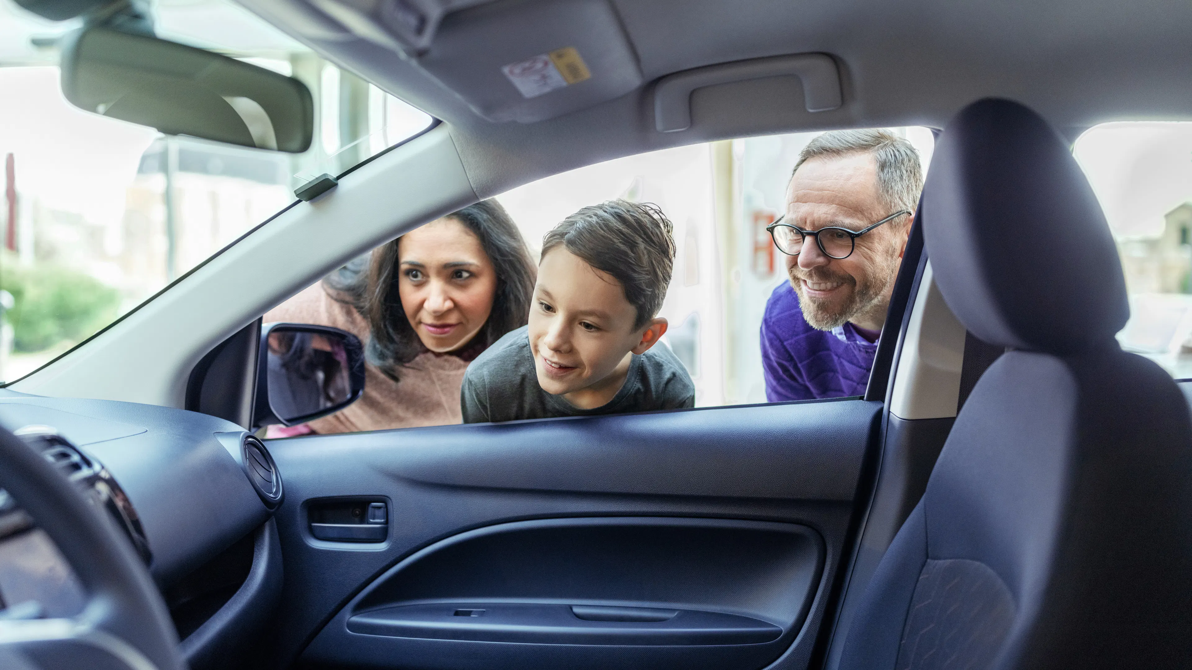 Eine Frau, ein Mann und ein Kind schauen gemeinsam von außen durch die geöffnete Autoscheibe in den Innenraum eines Fahrzeugs.