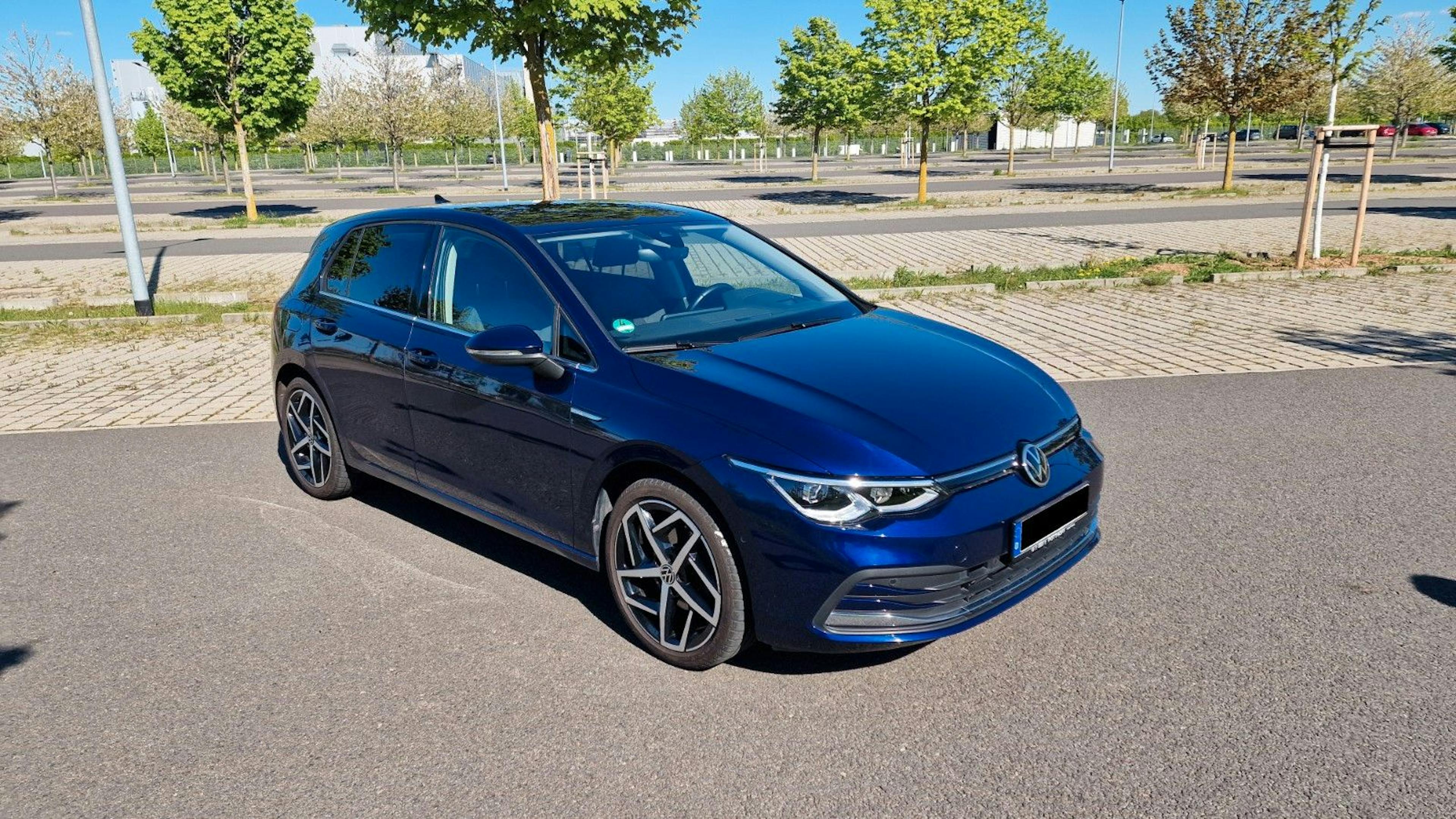 Dunkelblauer VW Golf 8 und sportlichen Leichtmetallfelgen, geparkt auf einem sonnigen, leeren Parkplatz mit Bäumen im Hintergrund.