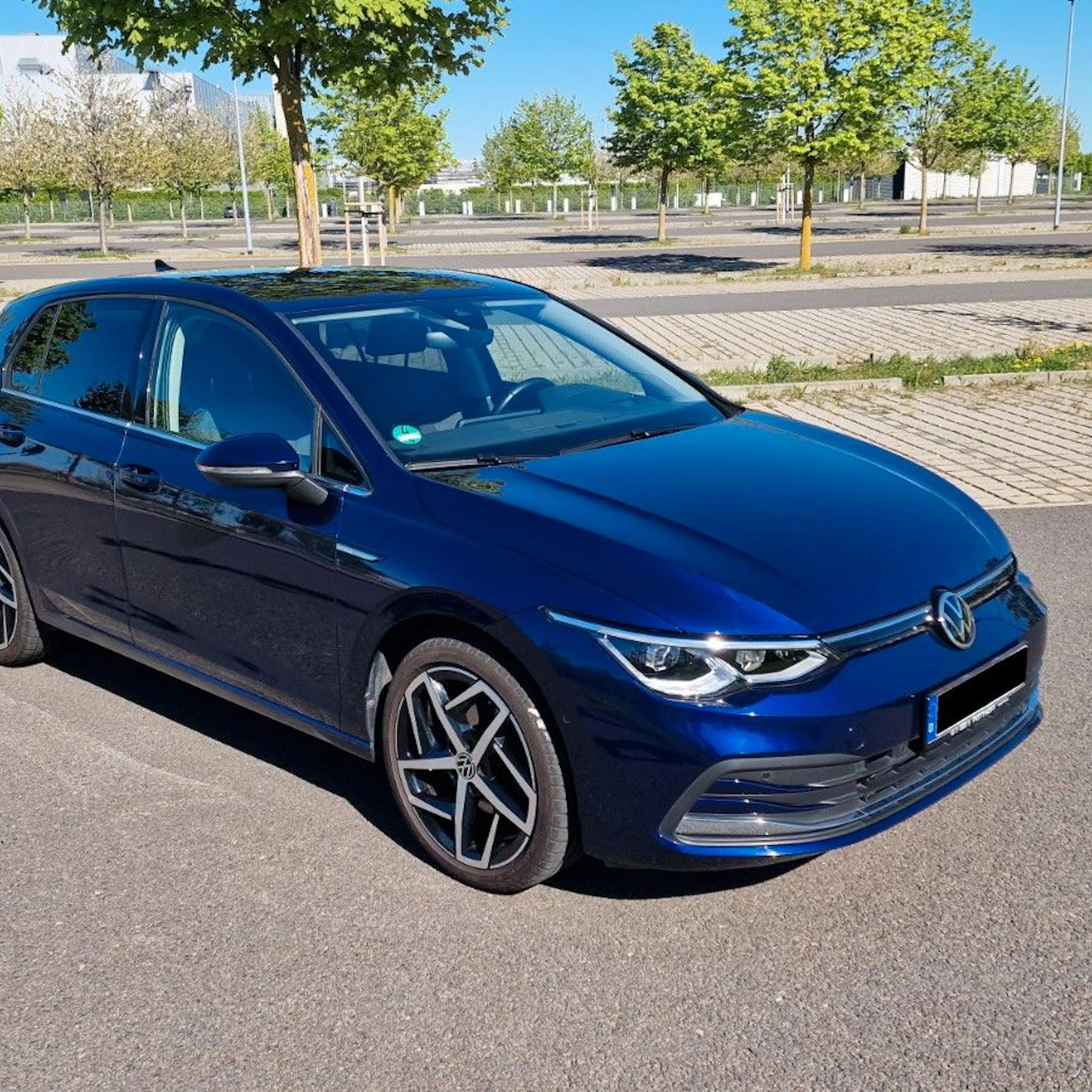 Dunkelblauer VW Golf 8 und sportlichen Leichtmetallfelgen, geparkt auf einem sonnigen, leeren Parkplatz mit Bäumen im Hintergrund.